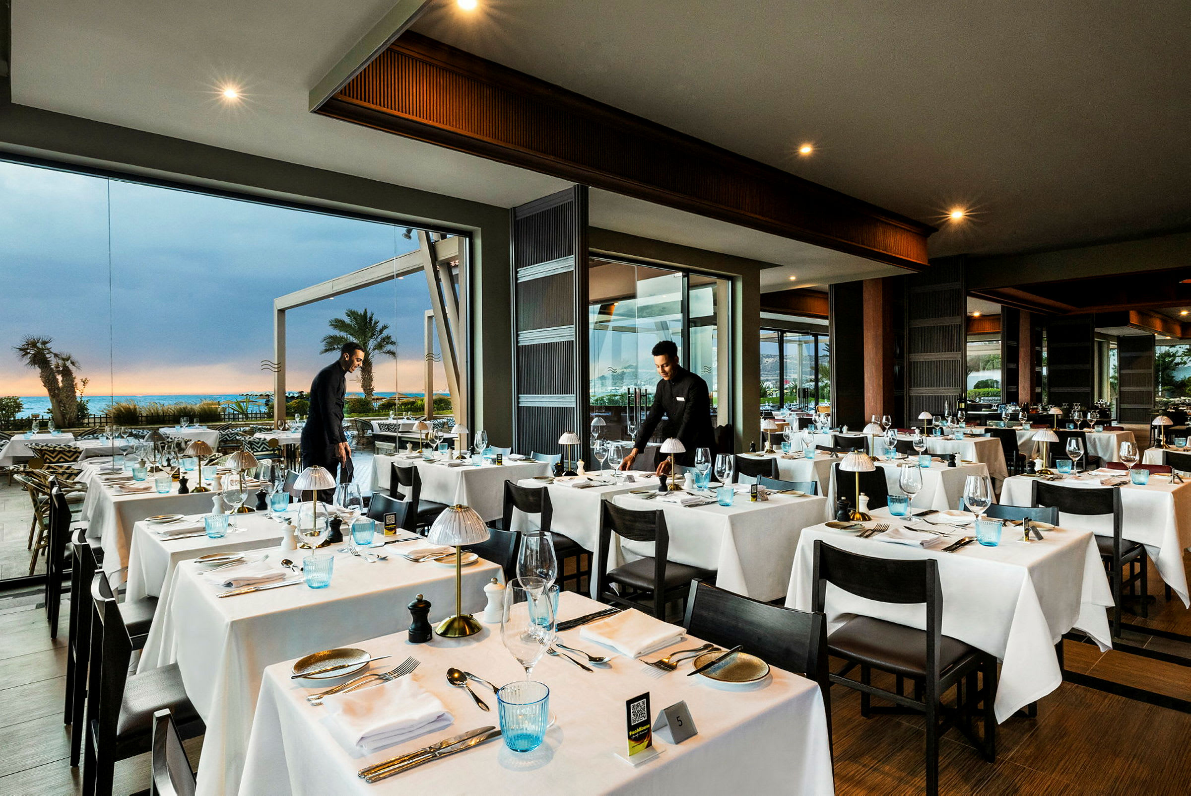 Interiörbild av en restaurang med dukade bord och stora glasfönster som visar utsikten mot havet och palmer. (AI-genererad text)