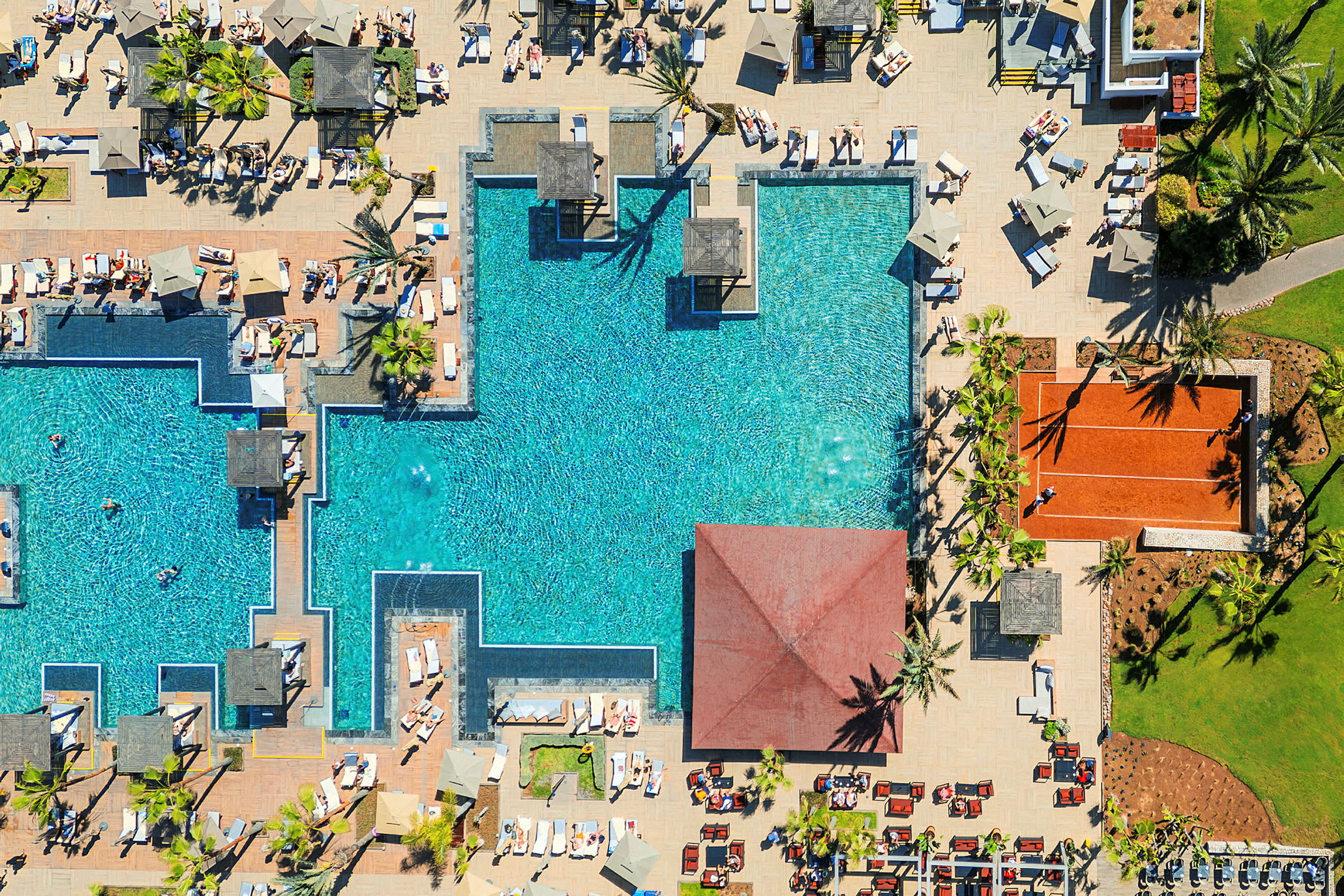 Flygfoto av en pool, solstolar, parasoller och en tennisbana. (AI-genererad text)