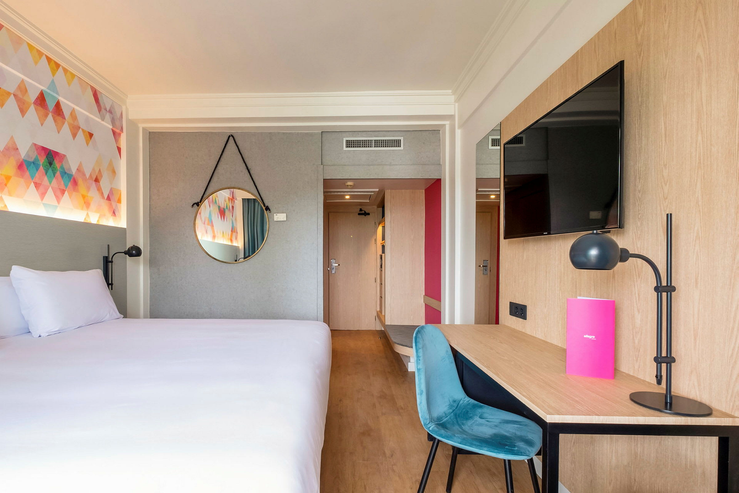 Ett hotellrum med en säng, ett skrivbord med stol och lampa, samt en vägg-tv. (AI-genererad text)