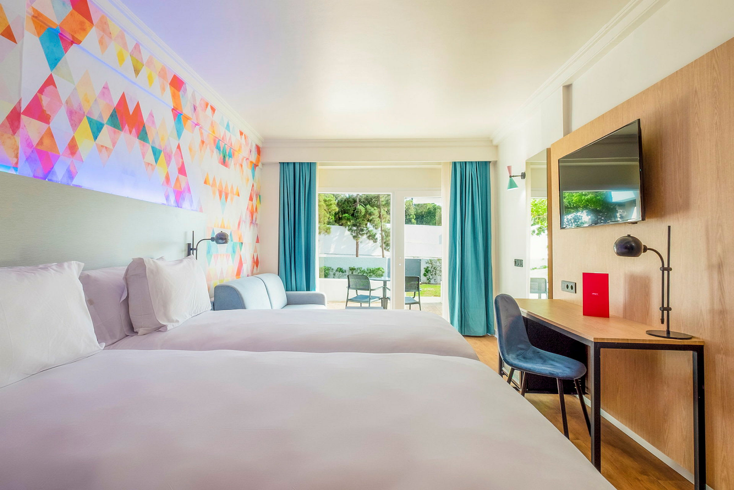 Ett hotellrum med två sängar, en färgglad väggdesign, sittgrupp, skrivbord och tillgång till en terass med stolar. (AI-genererad text)