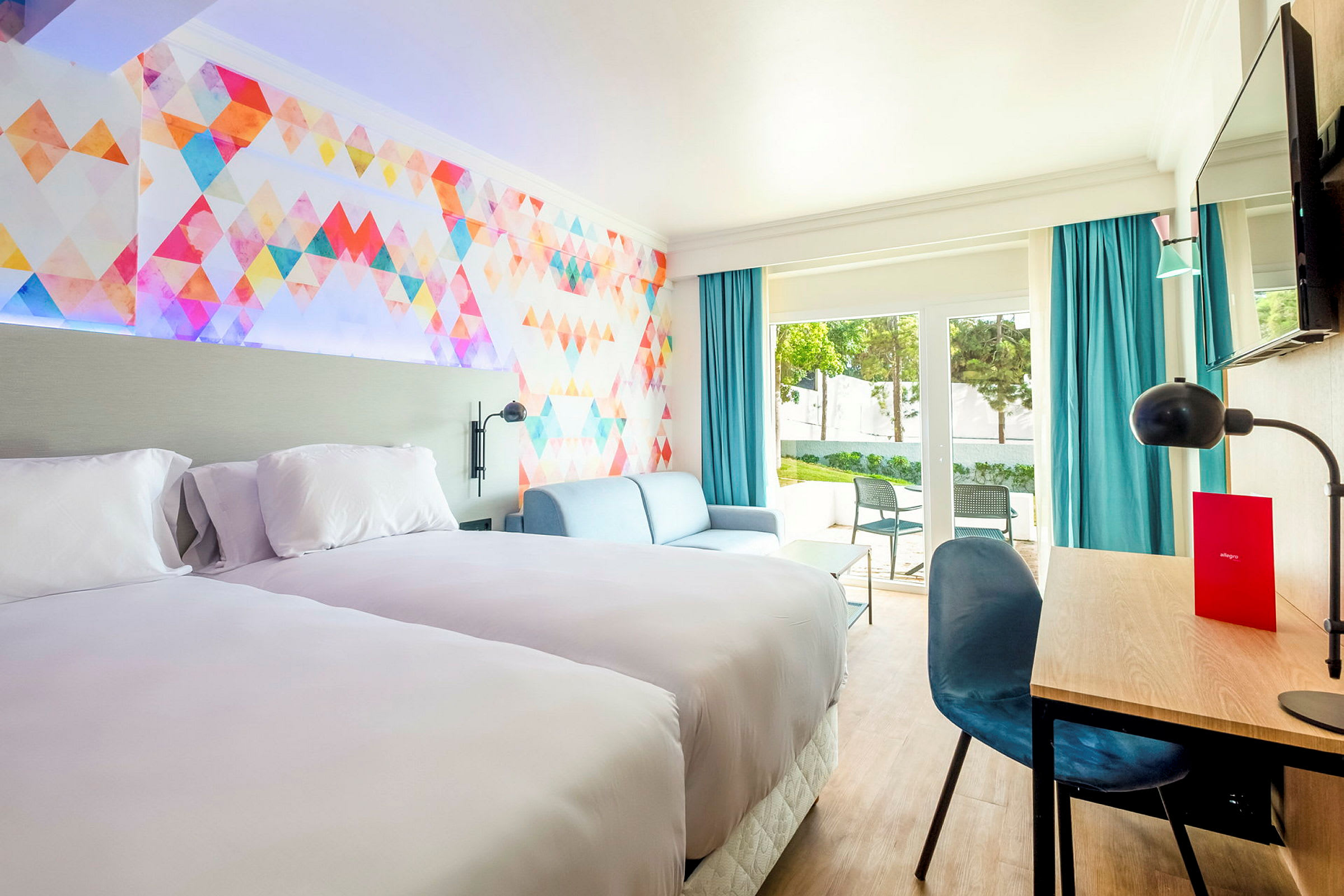 Hotellrum med två sängar, soffa och skrivbord. Färgglad väggdesign och stora fönster. (AI-genererad text)