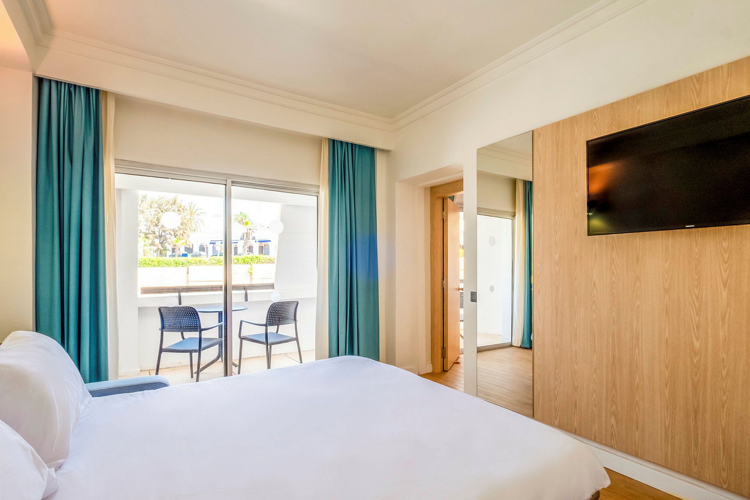 Ett hotellrum med en säng, en väggmonterad TV och en balkong med två stolar. (AI-genererad text)