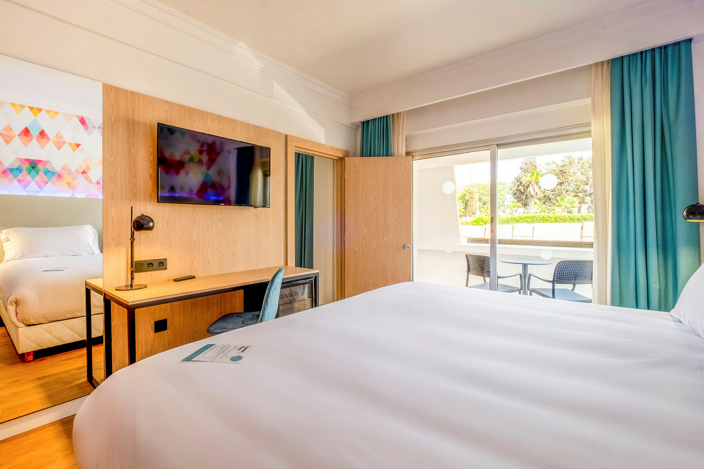 Ett hotellrum med en dubbelsäng, skrivbord, stol och en TV på väggen. Det finns en balkong med utsikt. (AI-genererad text)