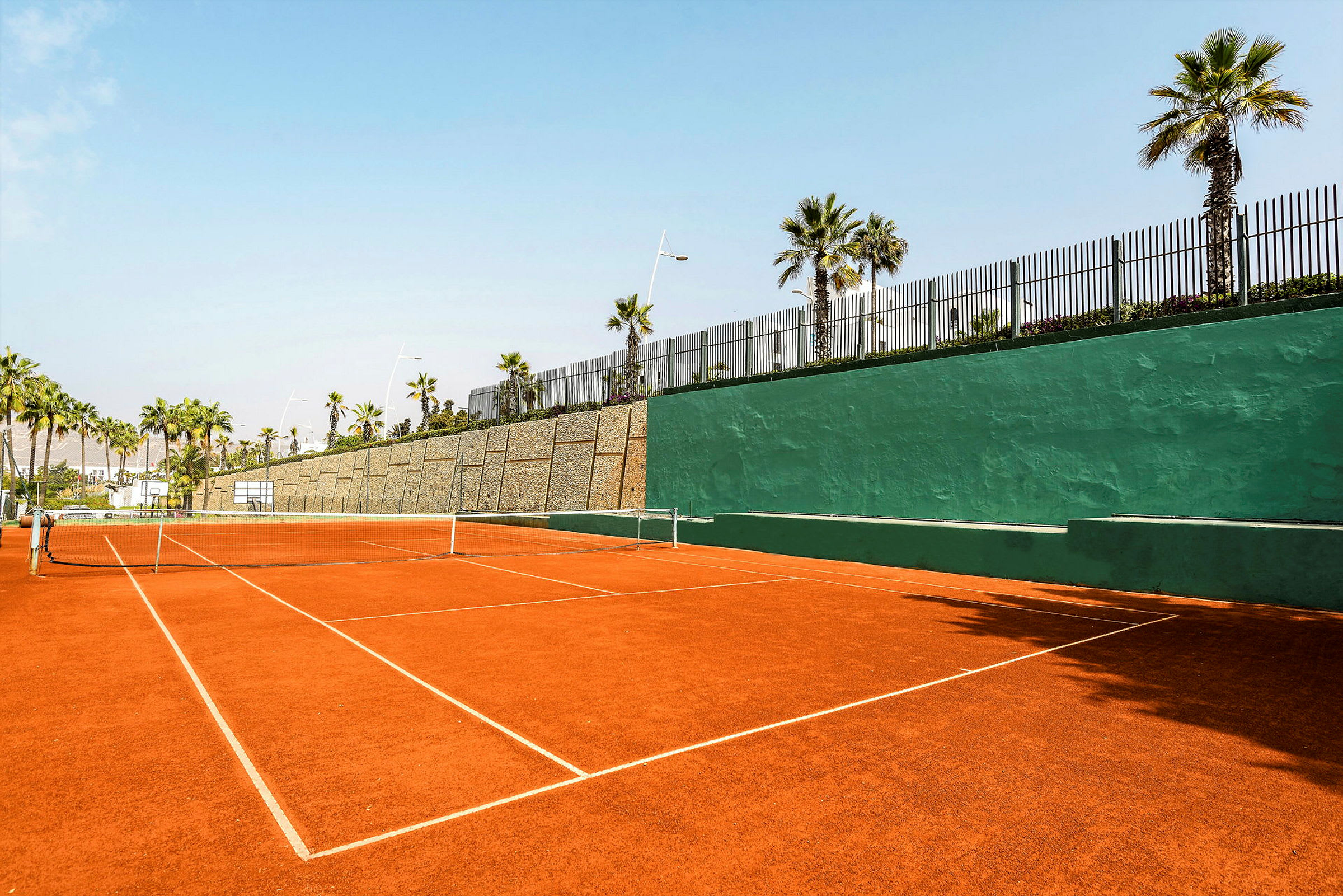 En tennisbana med röd grus och ett grönt plank i bakgrunden, omgiven av palmer. (AI-genererad text)