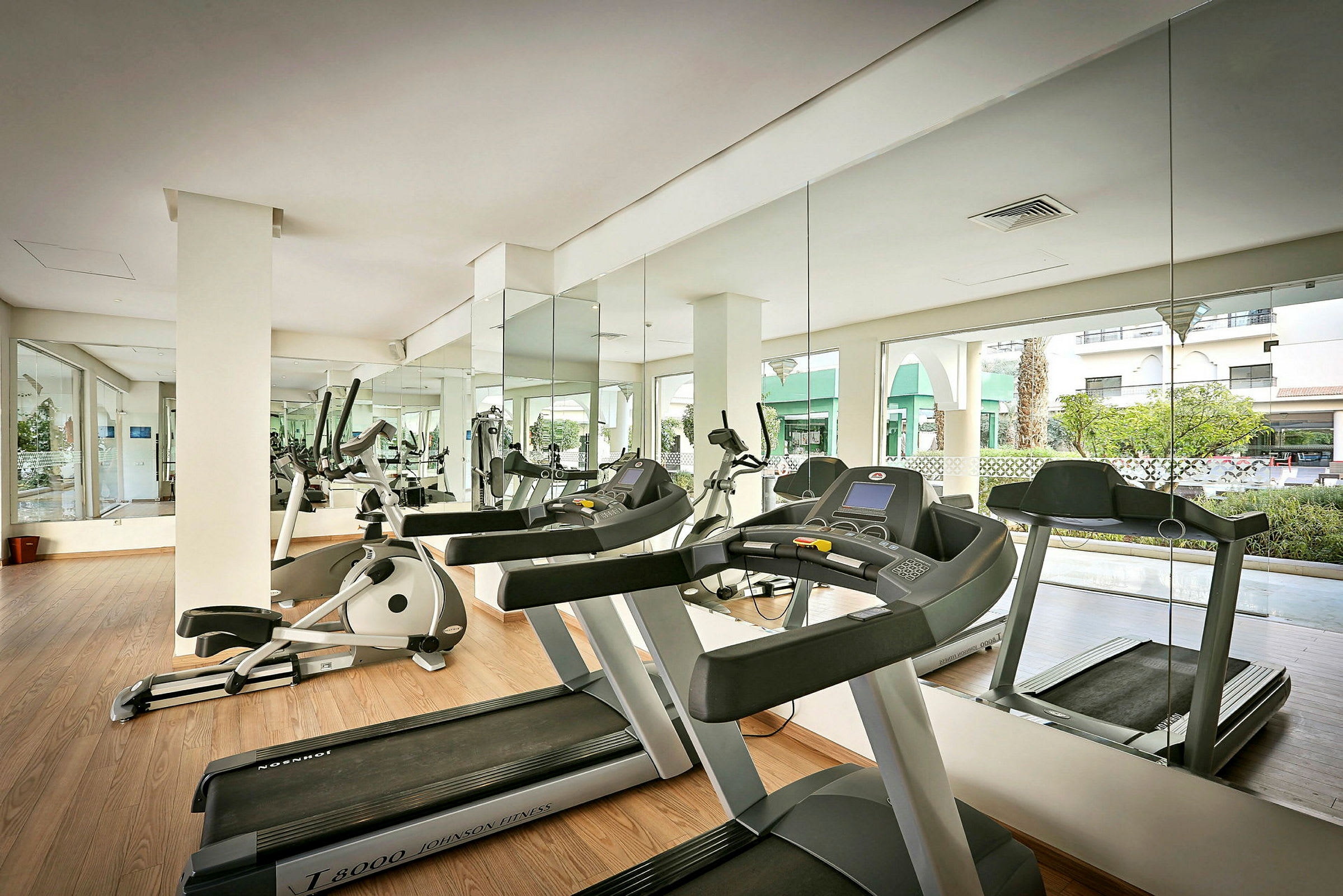 Ett modernt gym med löpband och träningsutrustning, speglar och fönster ut mot grönskande omgivningar. (AI-genererad text)