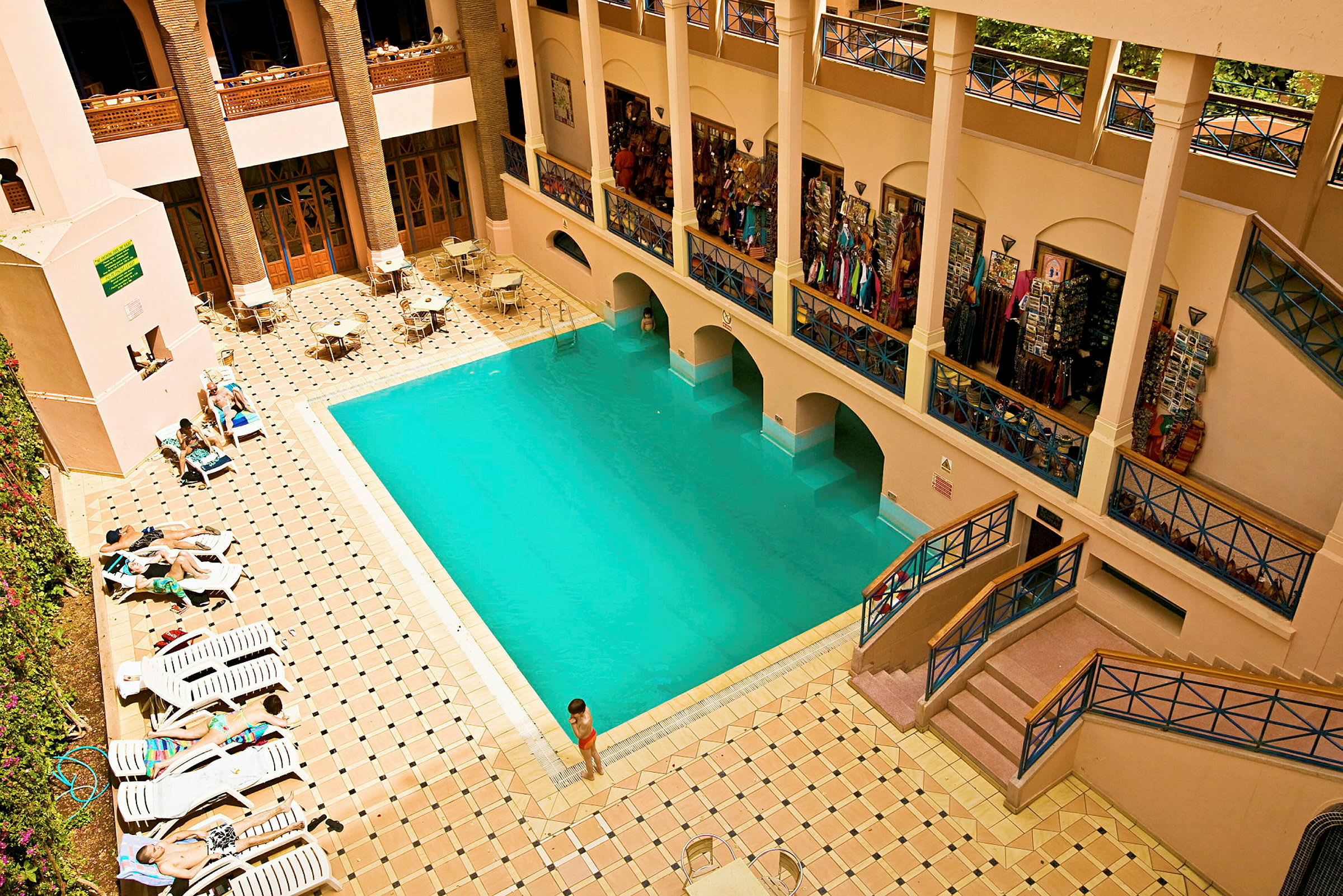 Ett inomhusområde med en swimmingpool omgiven av solstolar och bord under en balkong med affärer. (AI-genererad text)