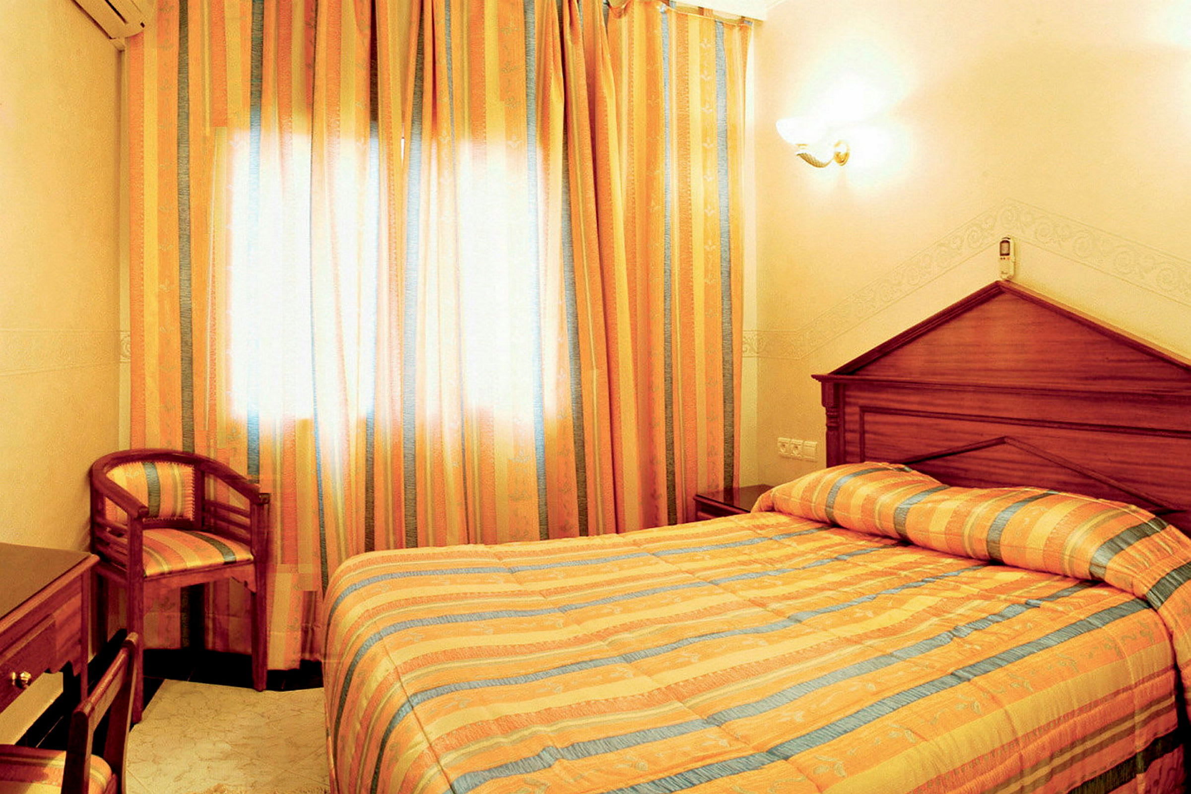 Ett hotellrum med en dubbelsäng klädd med randig sängklädsel i ljusa färger, en stol och ett skrivbord. Rummet har lysande gardiner och ljusa väggar. (AI-genererad text)