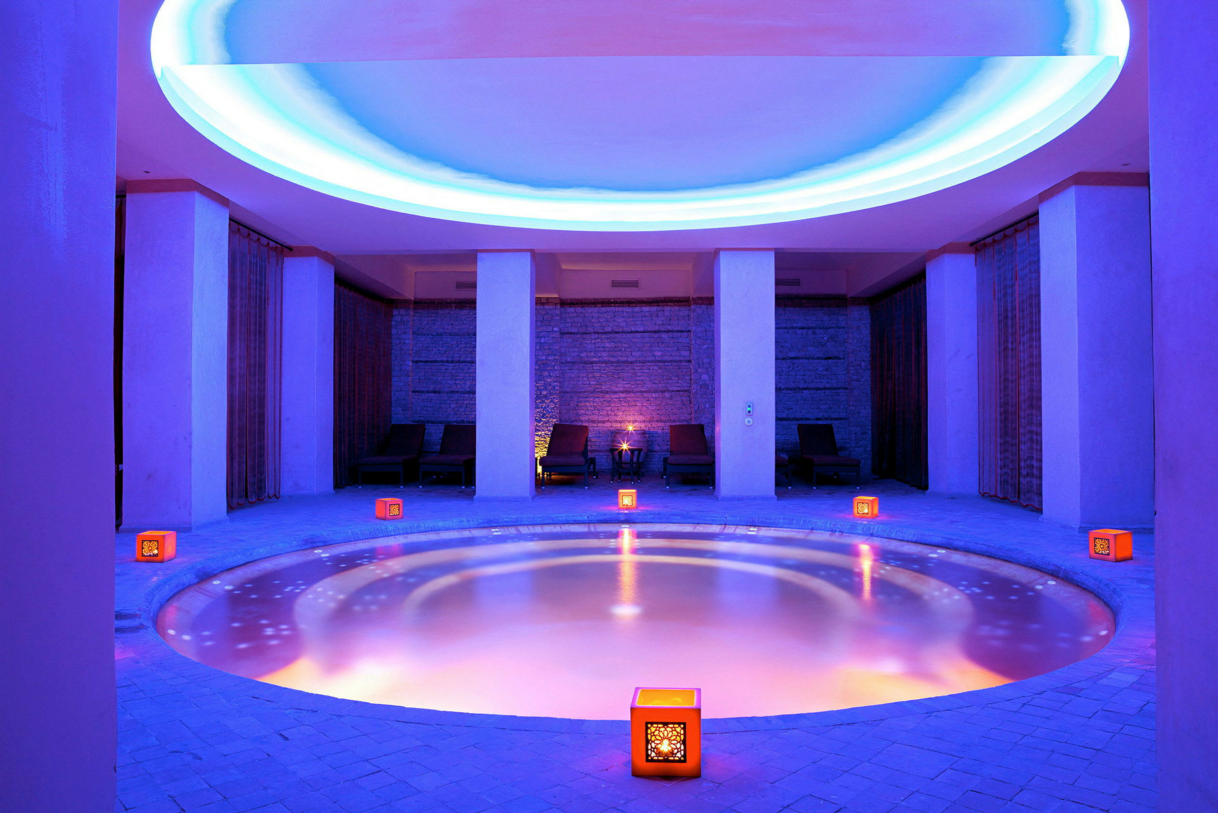 Ett blått upplyst spa med en rund pool omgiven av dekorativa ljus och vilstolar. (AI-genererad text)