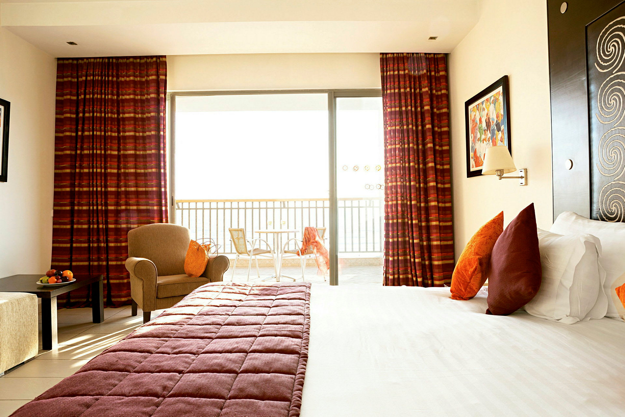 Ett hotellrum med en säng utrustad med kuddar och bäddsett i varma toner. Ett fönster med draperier och en balkong med bord och stolar. (AI-genererad text)