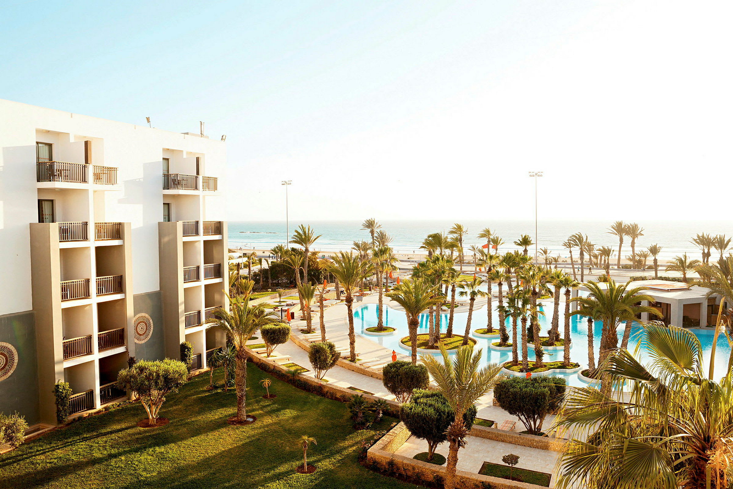 Hotellets fasad med balkonger och en vy över grönskande trädgårdar och en pool, samt havet i bakgrunden. (AI-genererad text)
