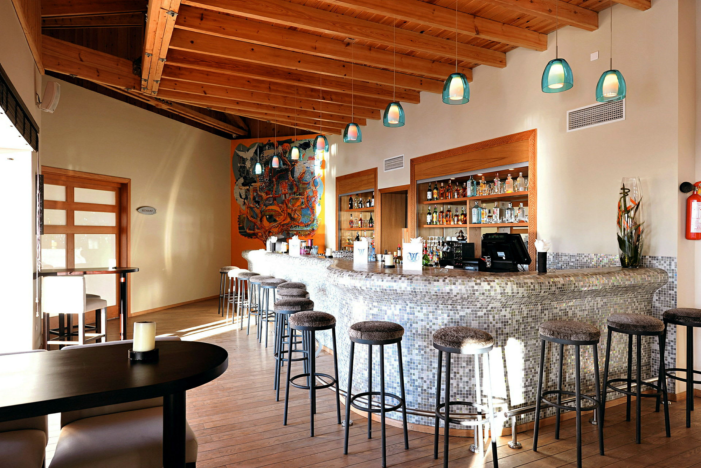 En modern bar med höga stolar och en bardisk med mosaik. Taket har träbjälkar och väggarna pryds av konstverk och hyllor med olika flaskor. (AI-genererad text)