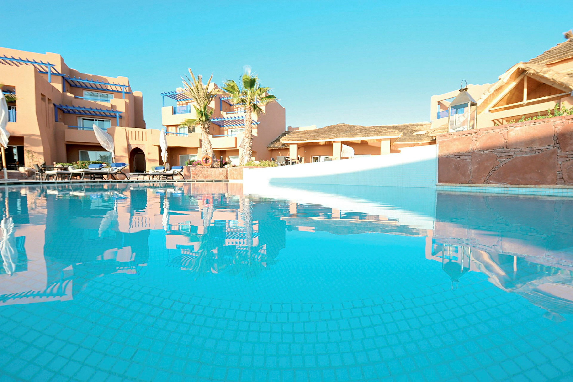 Bild av en pool omgiven av byggnader och palmer under en klar himmel. (AI-genererad text)