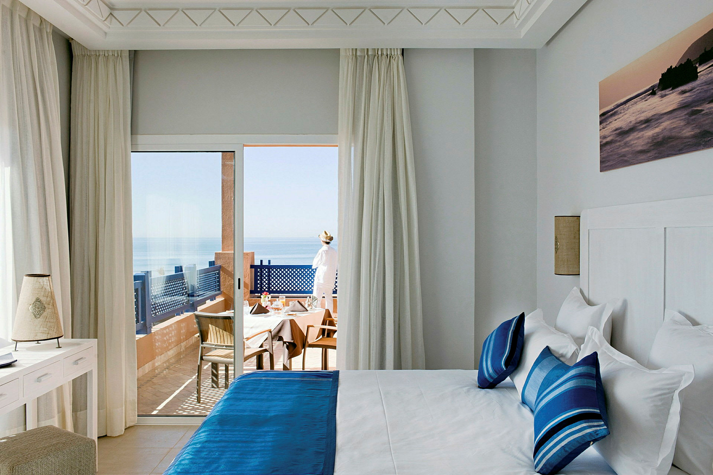 Ett hotellrum med en dubbelsäng med blå sängkläder och kuddar. Ett stort glasfönster visar en balkong med stolar och bord samt en utsikt över havet. (AI-genererad text)