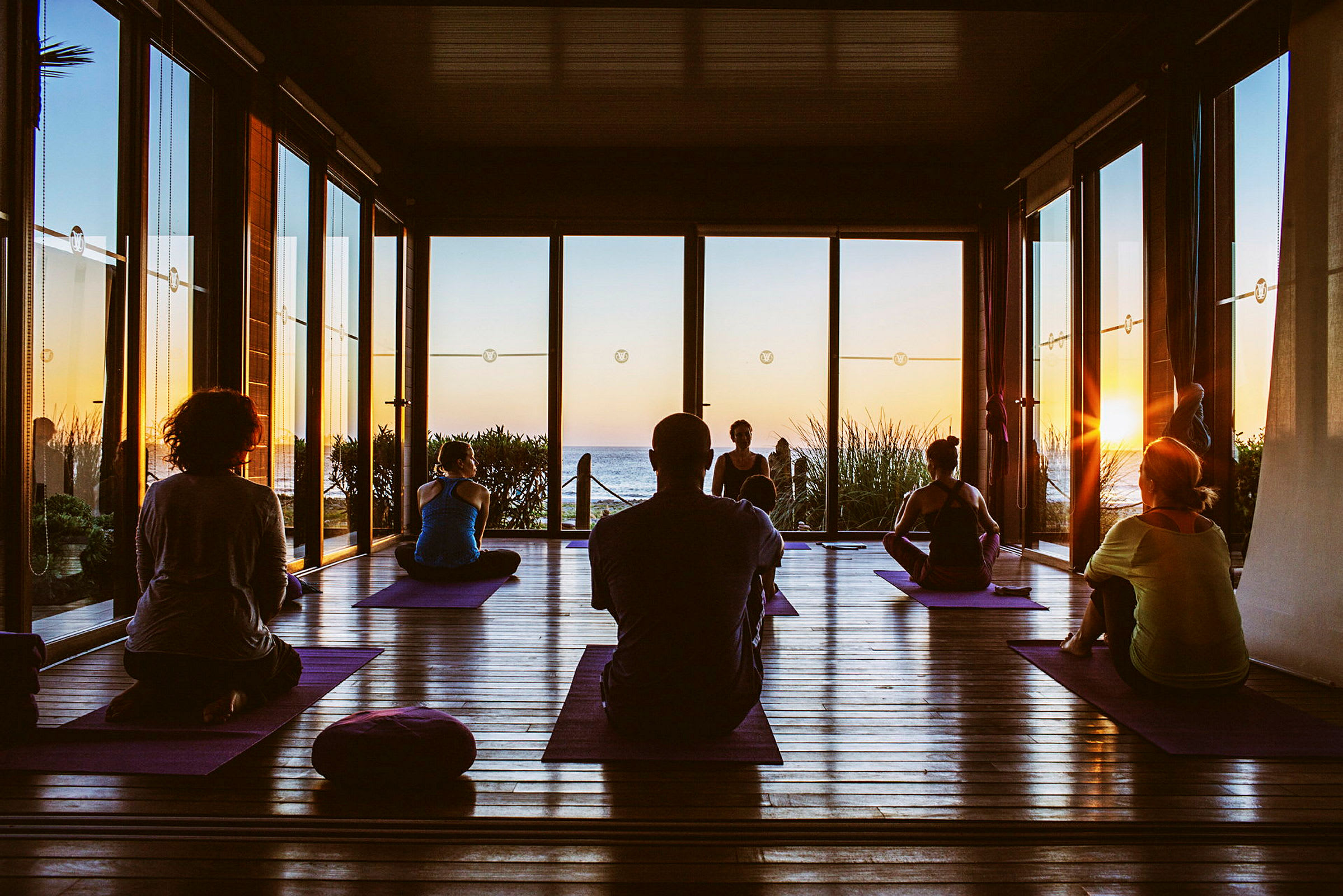 Personer som utövar yoga på mattor i en sal med utsikt över havet vid solnedgången. (AI-genererad text)