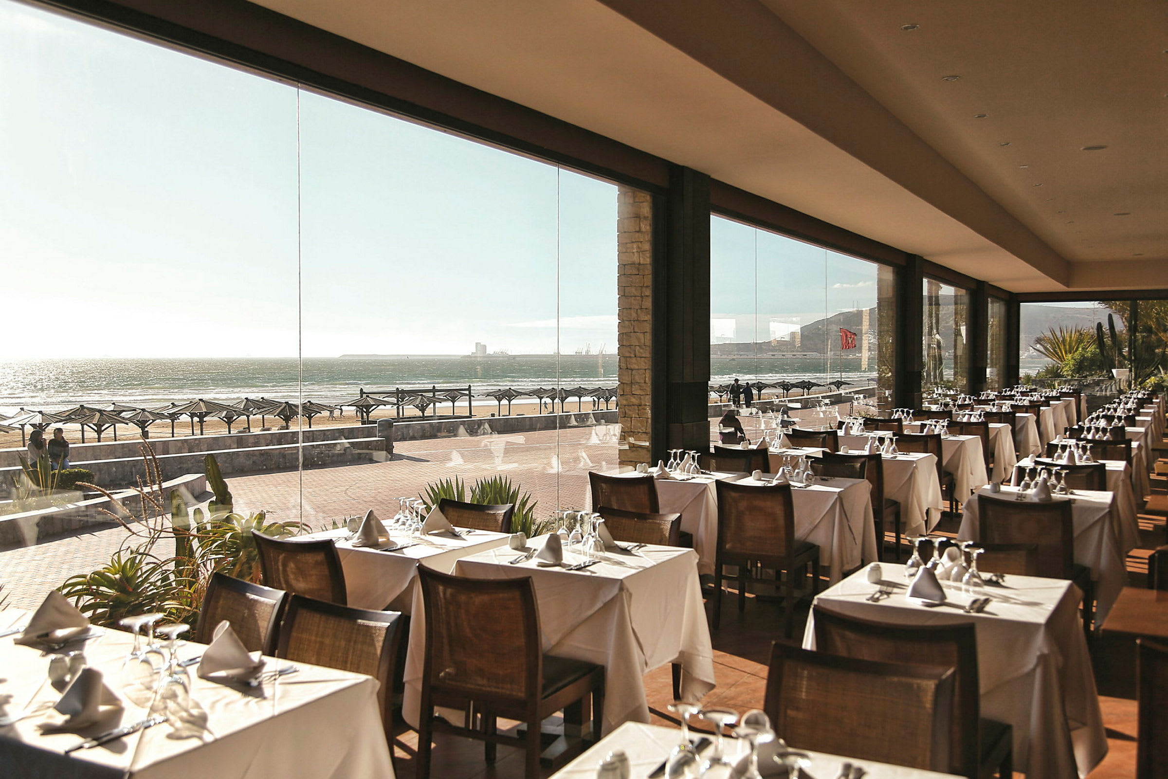 Restaurang med uppdukat bord och glasväggar som har utsikt över stranden. (AI-genererad text)