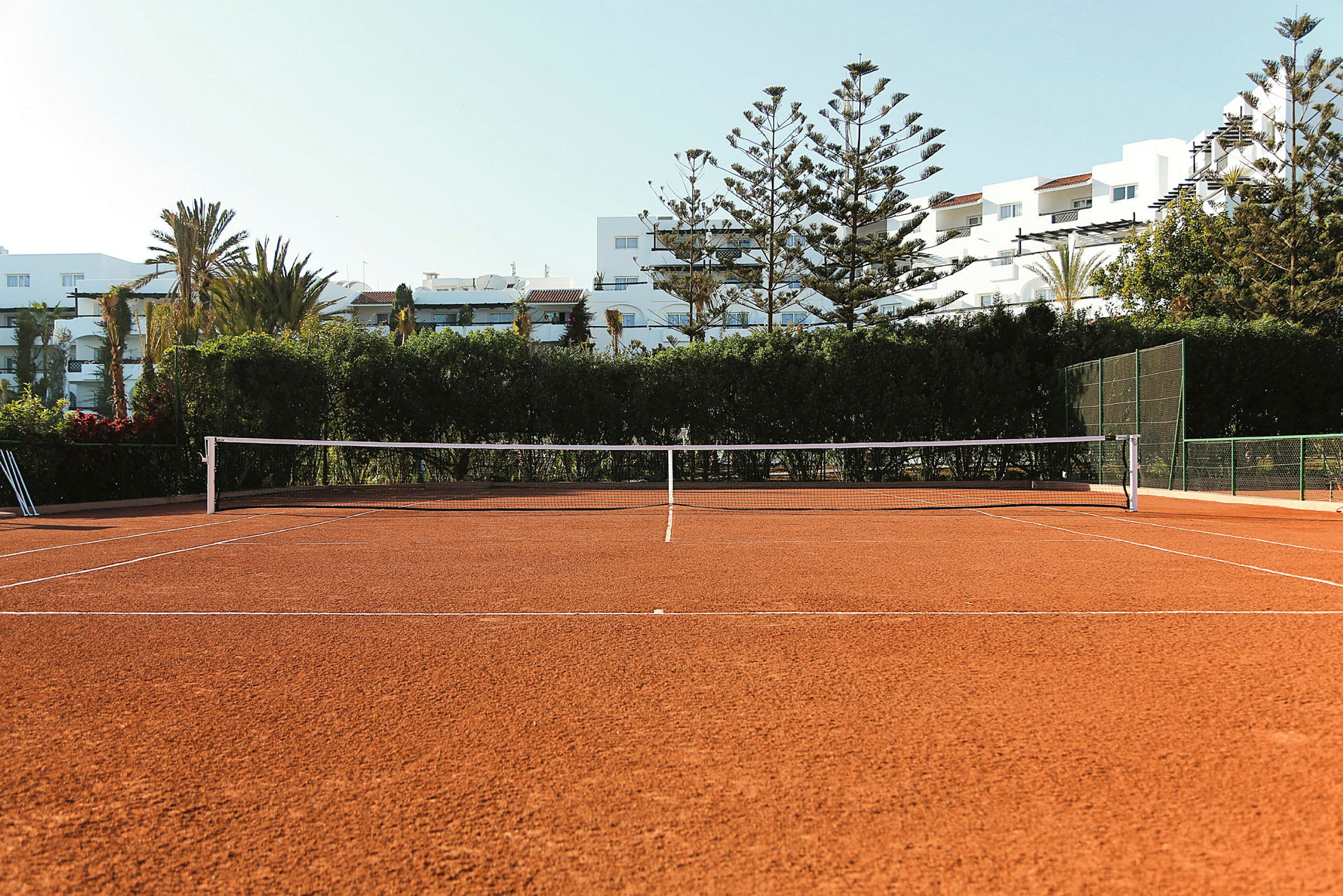 Bild av en tennisbana med grusunderlag, omgiven av träd och byggnader i bakgrunden. (AI-genererad text)