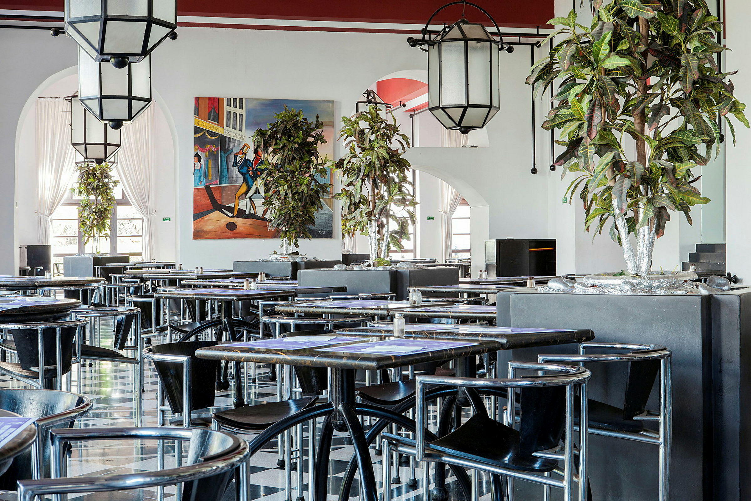 En inomhusrestaurang med bord och stolar. Detaljer som växter och en färgglad väggmålning är synliga. (AI-genererad text)