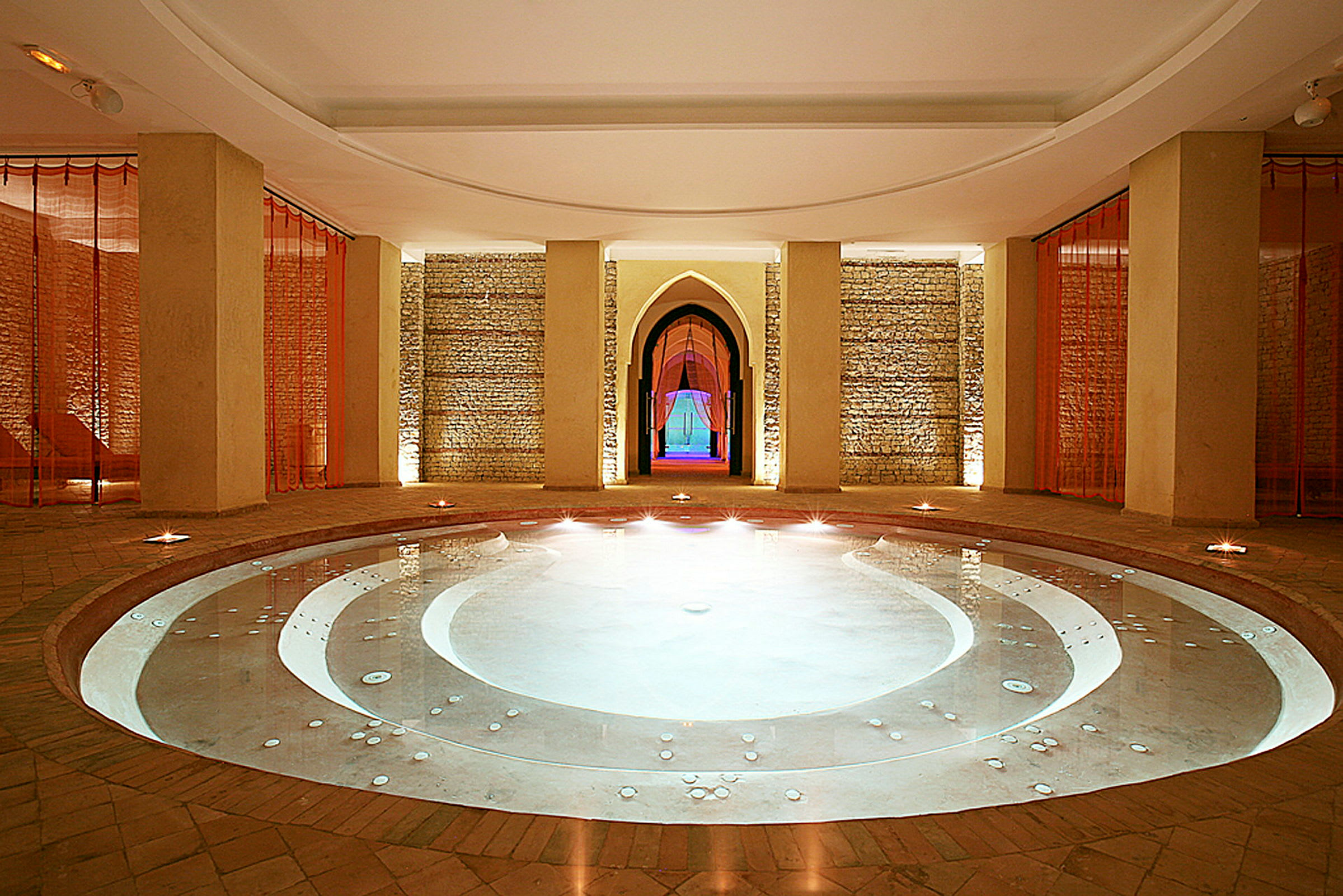 Ett elegant spa med en rund inomhuspool, omgiven av väggar med tegelstruktur och röda draperier. (AI-genererad text)