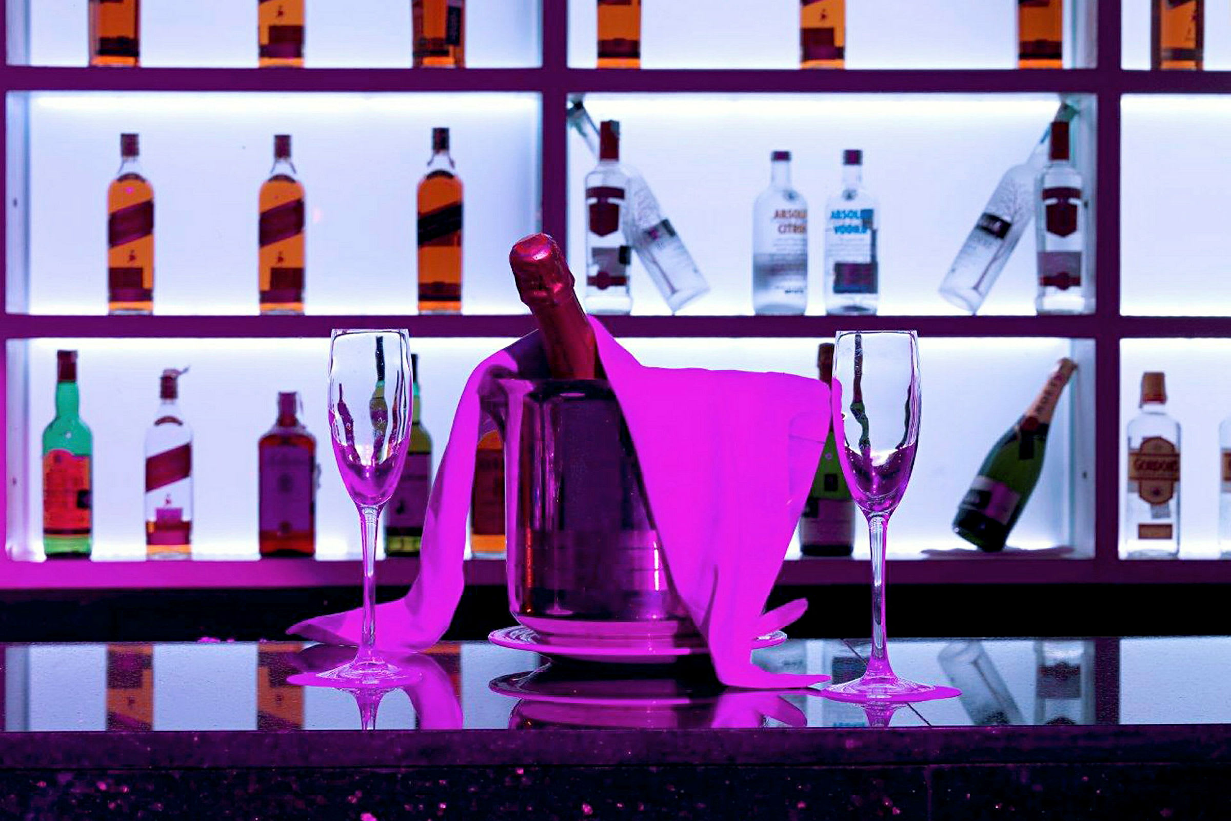 Ett bord med två tomma champagneglas och en champagneflaska i en ishink, omgiven av rosa belysning. I bakgrunden syns hyllor med flaskor. (AI-genererad text)