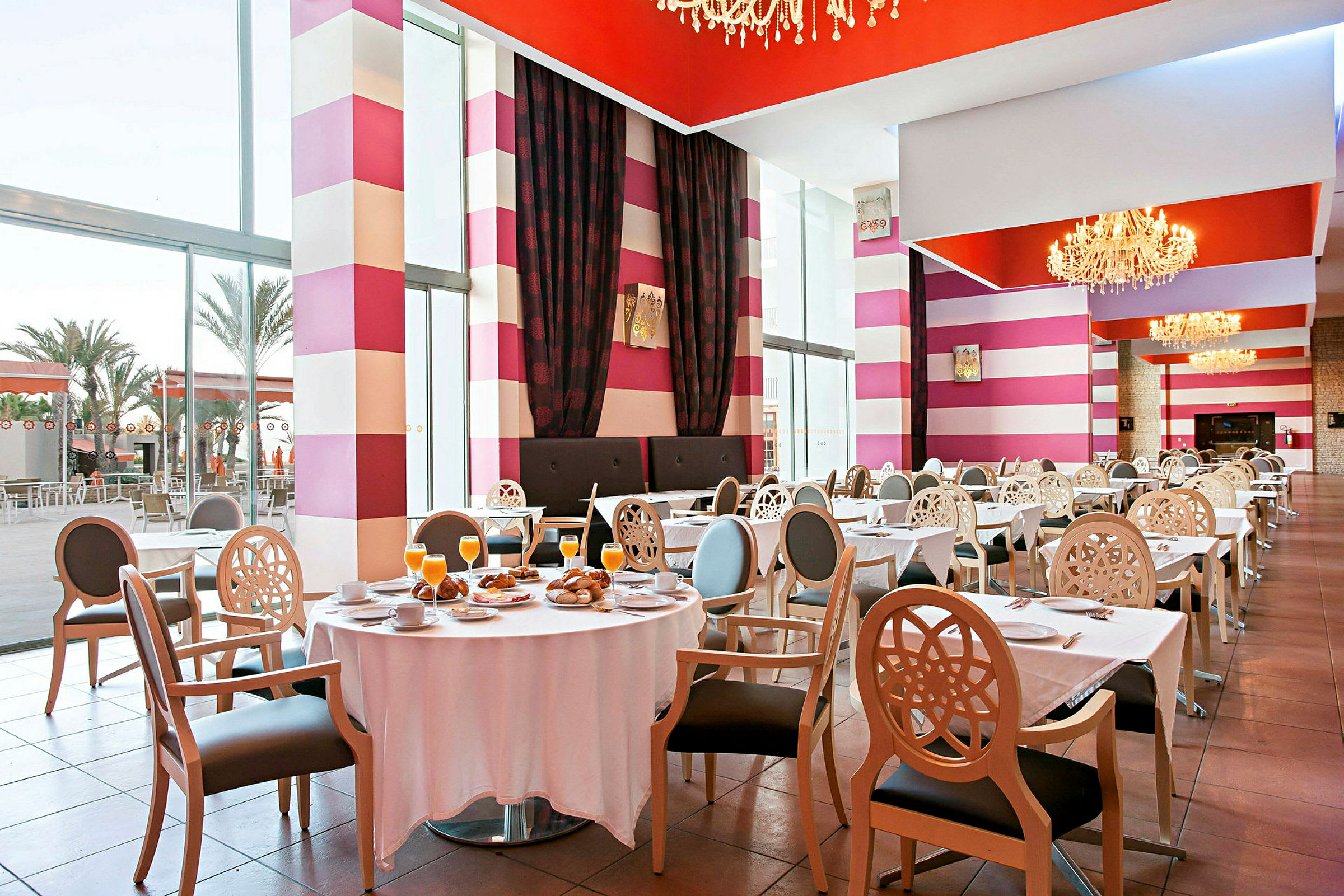 Ett välbelyst restaurangutrymme med dekorativa, färgglada väggar, dukade bord och stolar samt stora fönster som vetter ut mot ett utomhusområde. (AI-genererad text)