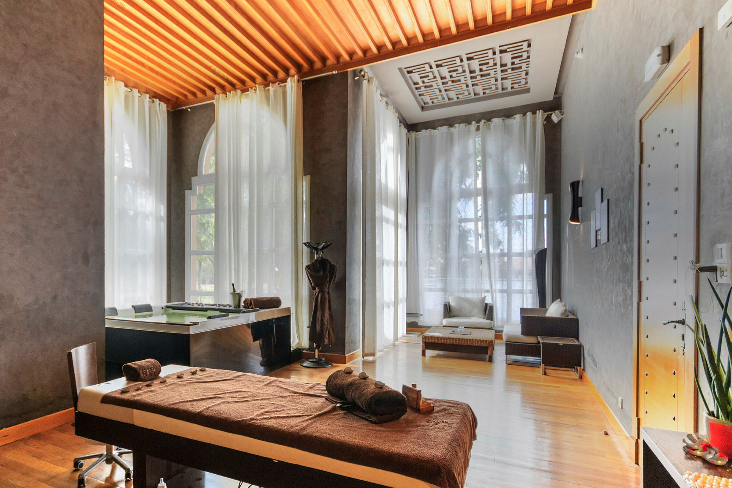 Ett spa- eller massageutrymme med en massagebänk, stolar och mjuka gardiner framför stora fönster. (AI-genererad text)