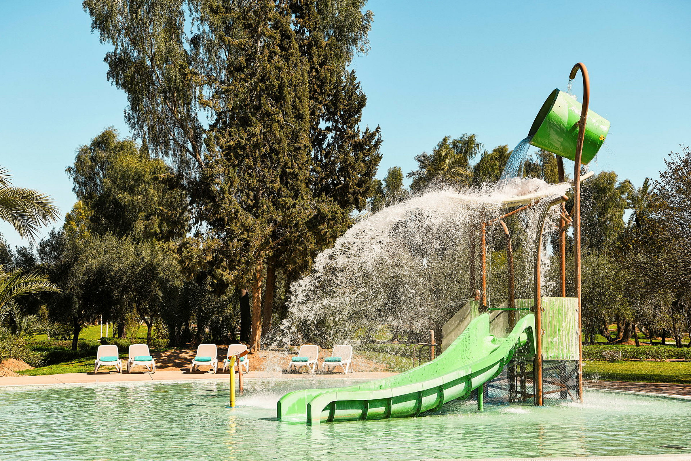 En lekanläggning vid en pool med vattenrutschbana och en stor grön hink som häller vatten. (AI-genererad text)
