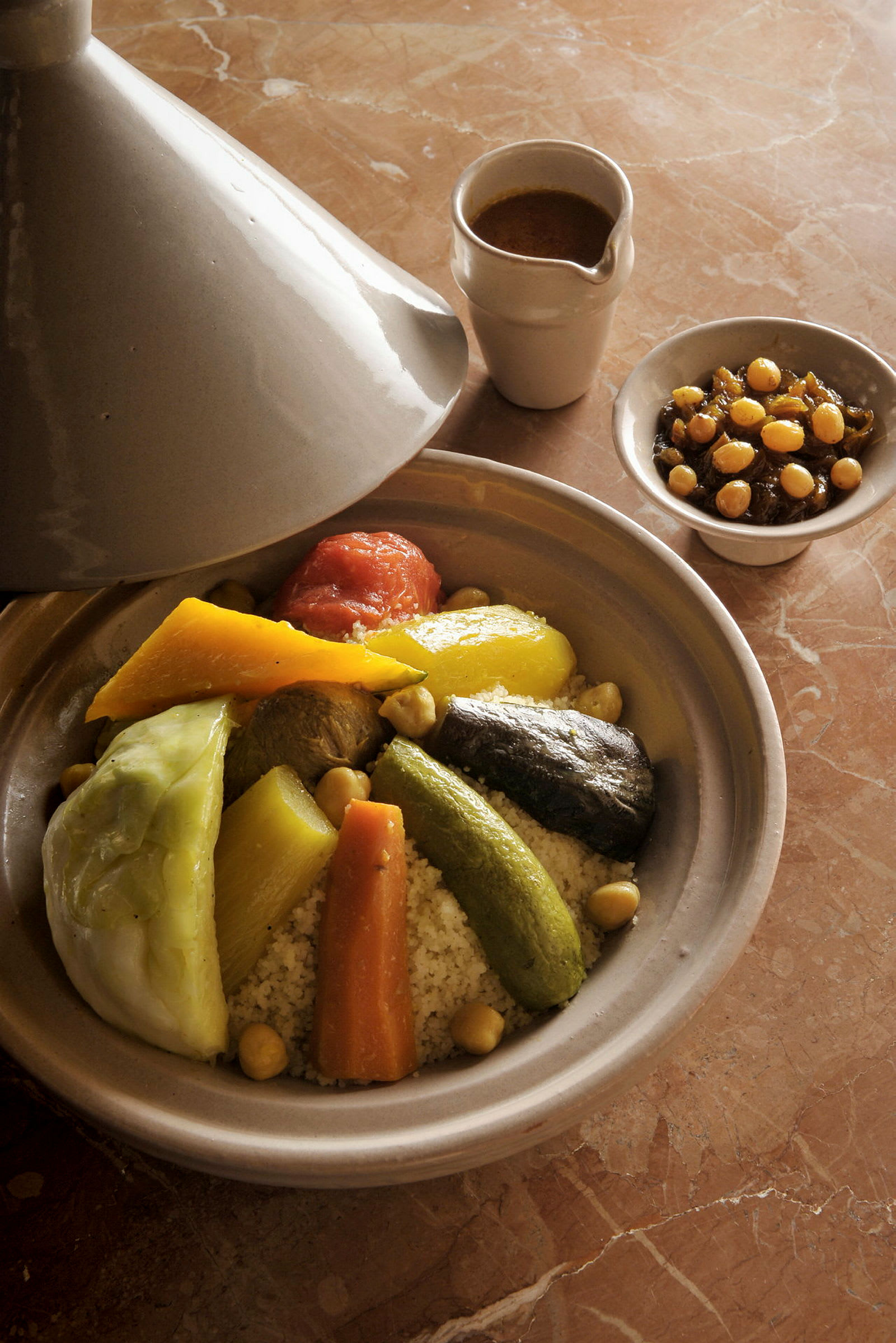 En traditionell tagine gryta med grönsaker och couscous, serveras med sås och en skål med kikärtor. På ett marmorbord. (AI-genererad text)