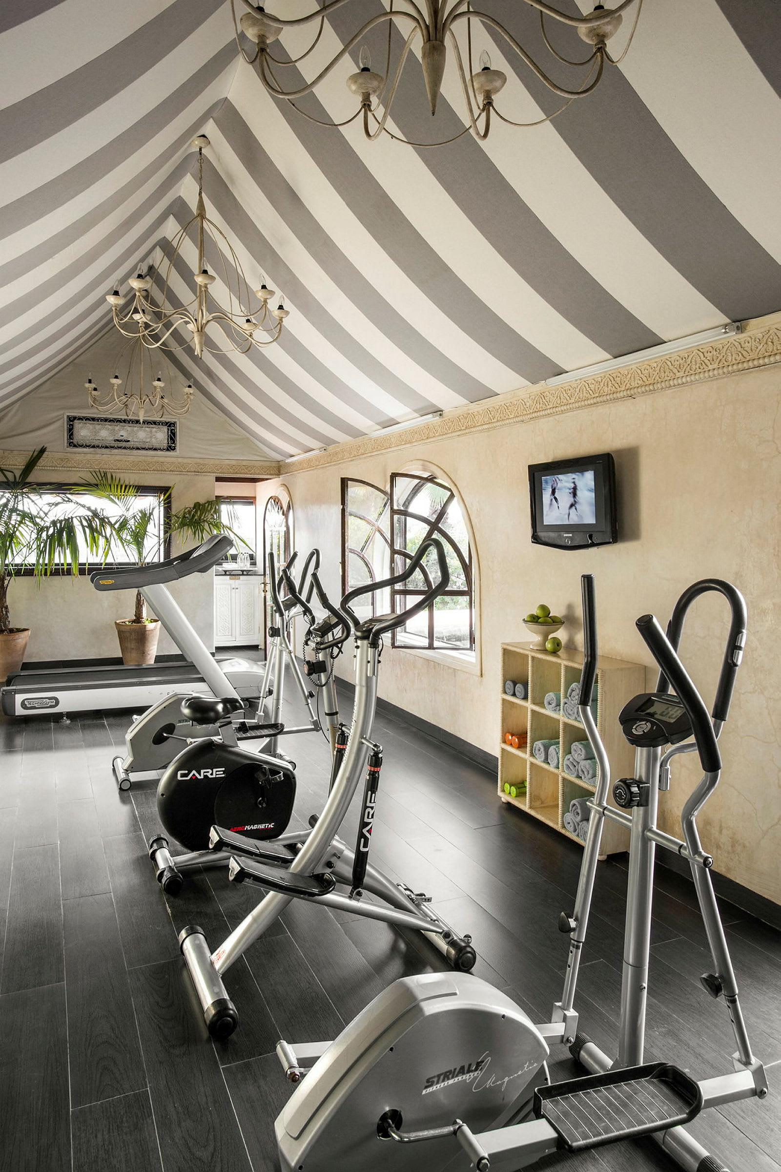 Ett gym med träningsutrustning, inklusive träningscyklar och löpband, i ett rum med randigt tak och stora fönster. (AI-genererad text)