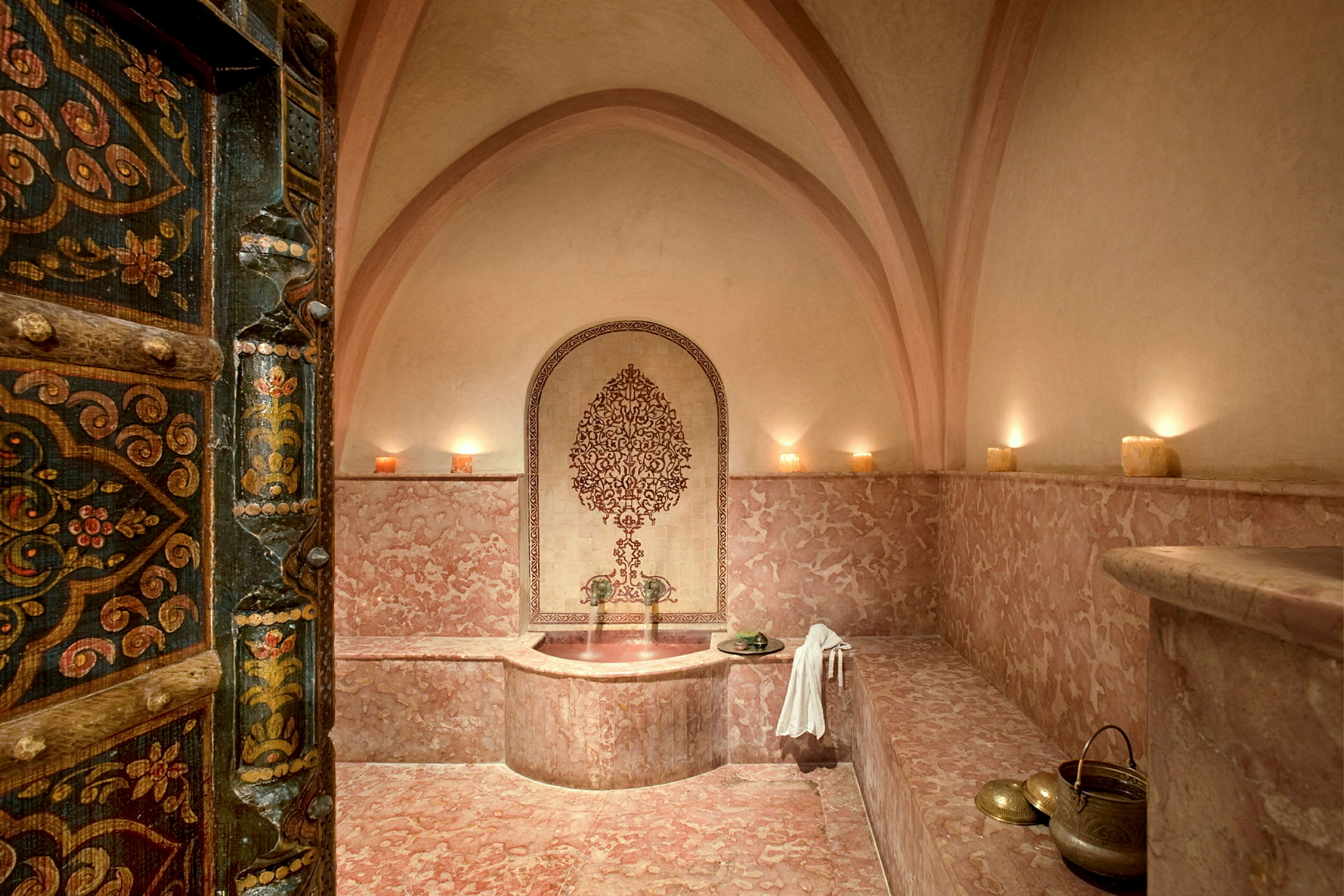 Ett traditionellt orientaliskt badrum med marmorväggar och valvad arkitektur. Dekorerade dörrar och stilfull belysning med ljus. (AI-genererad text)