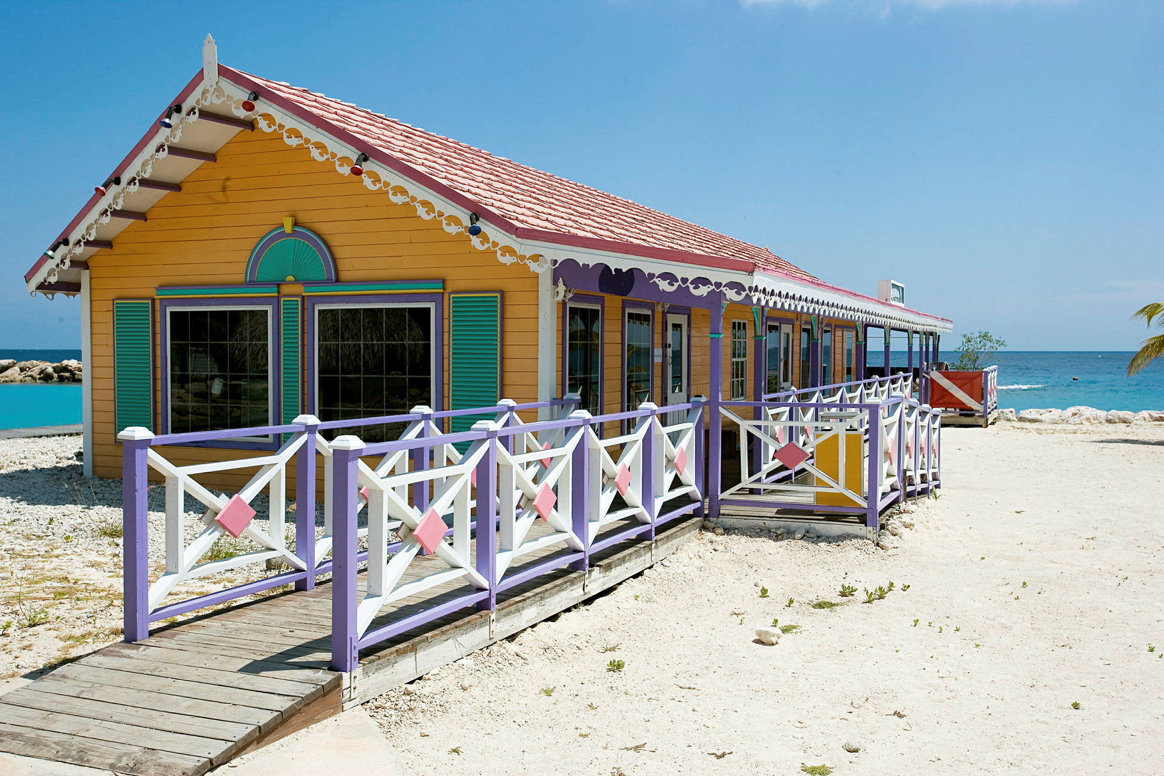 Ett färgglatt hus med dekorer och veranda, beläget vid en strand med klart blått vatten. (AI-genererad text)