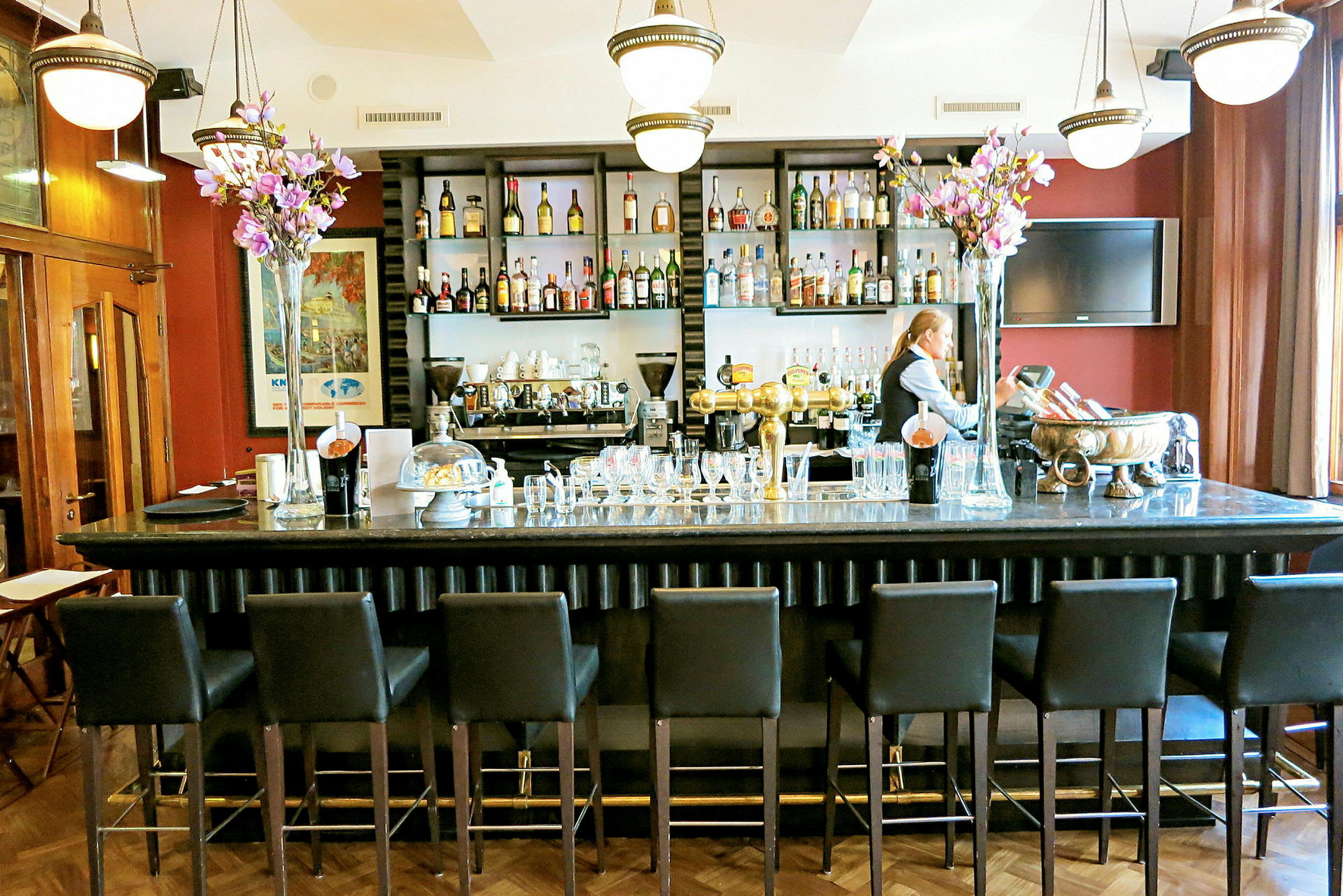 En bar med höga stolar framför en disk med glas och flaskor, bakom vilken en bartender står. (AI-genererad text)