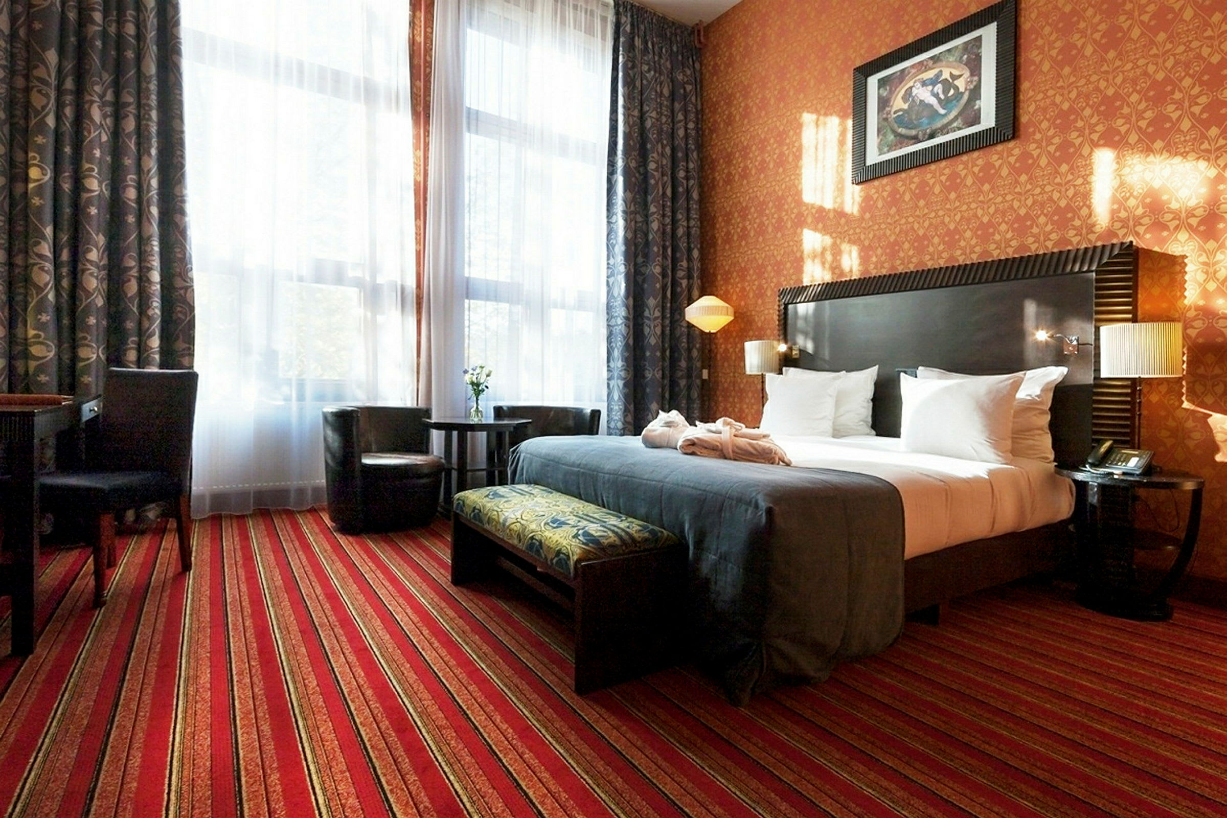 Ett hotellrum med färgglada tapeter och gardiner. Rummet har en dubbelsäng med vita sängkläder och ett mörkt överkast. Det finns en sittgrupp med två fåtöljer och ett bord samt ett skrivbord med stol. (AI-genererad text)