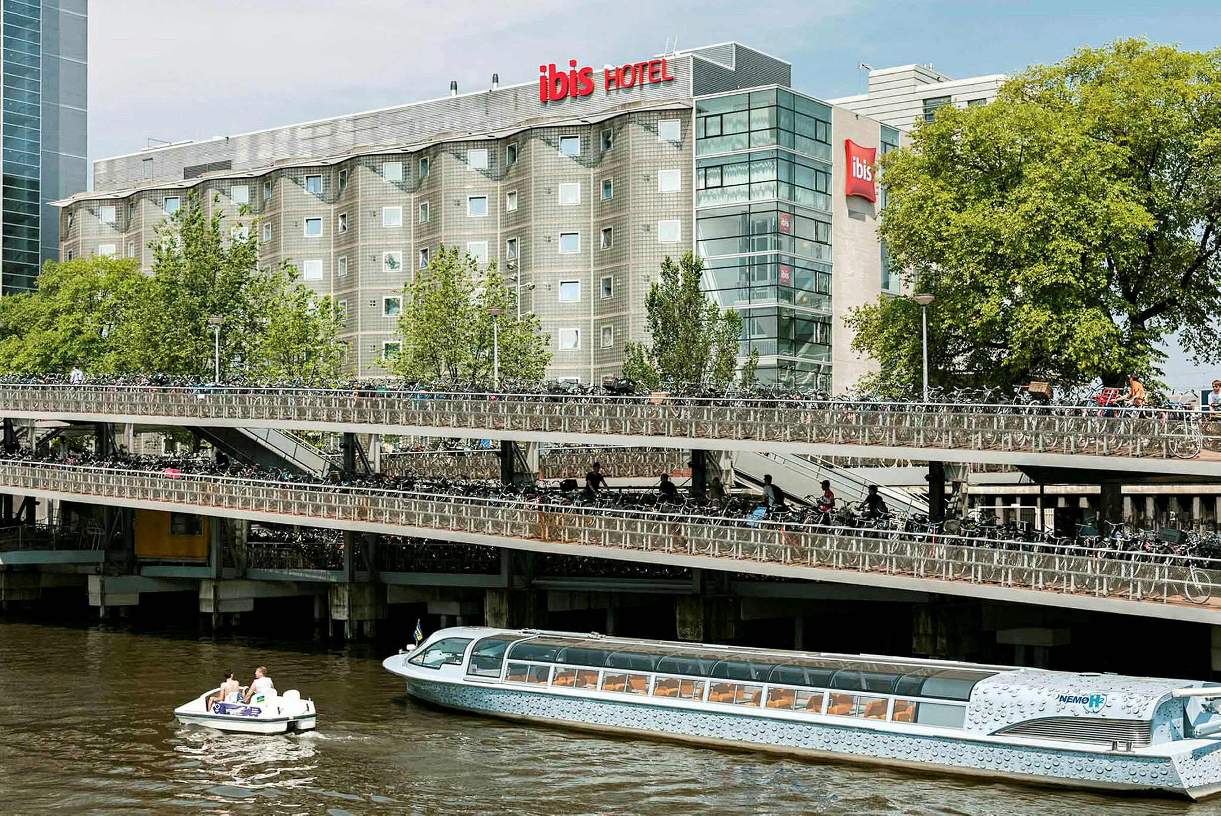 Bild av ett trevånings parkeringshus för cyklar bredvid en flod, med ett hotell i bakgrunden och båtar på vattnet. (AI-genererad text)