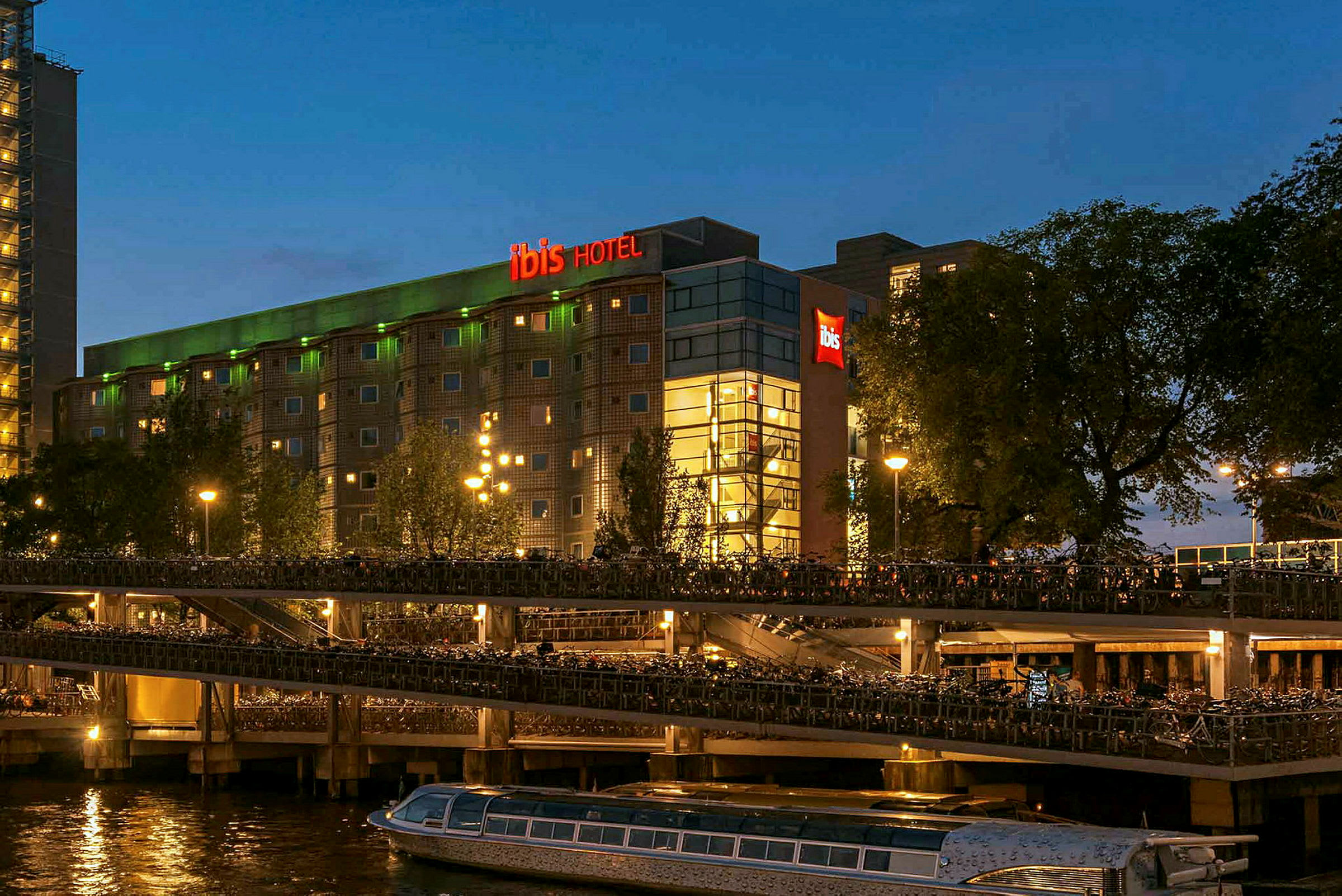 Hotellbyggnad kvällstid med neonskylt i rött och grönt, vatten och husbåtar i förgrunden. (AI-genererad text)