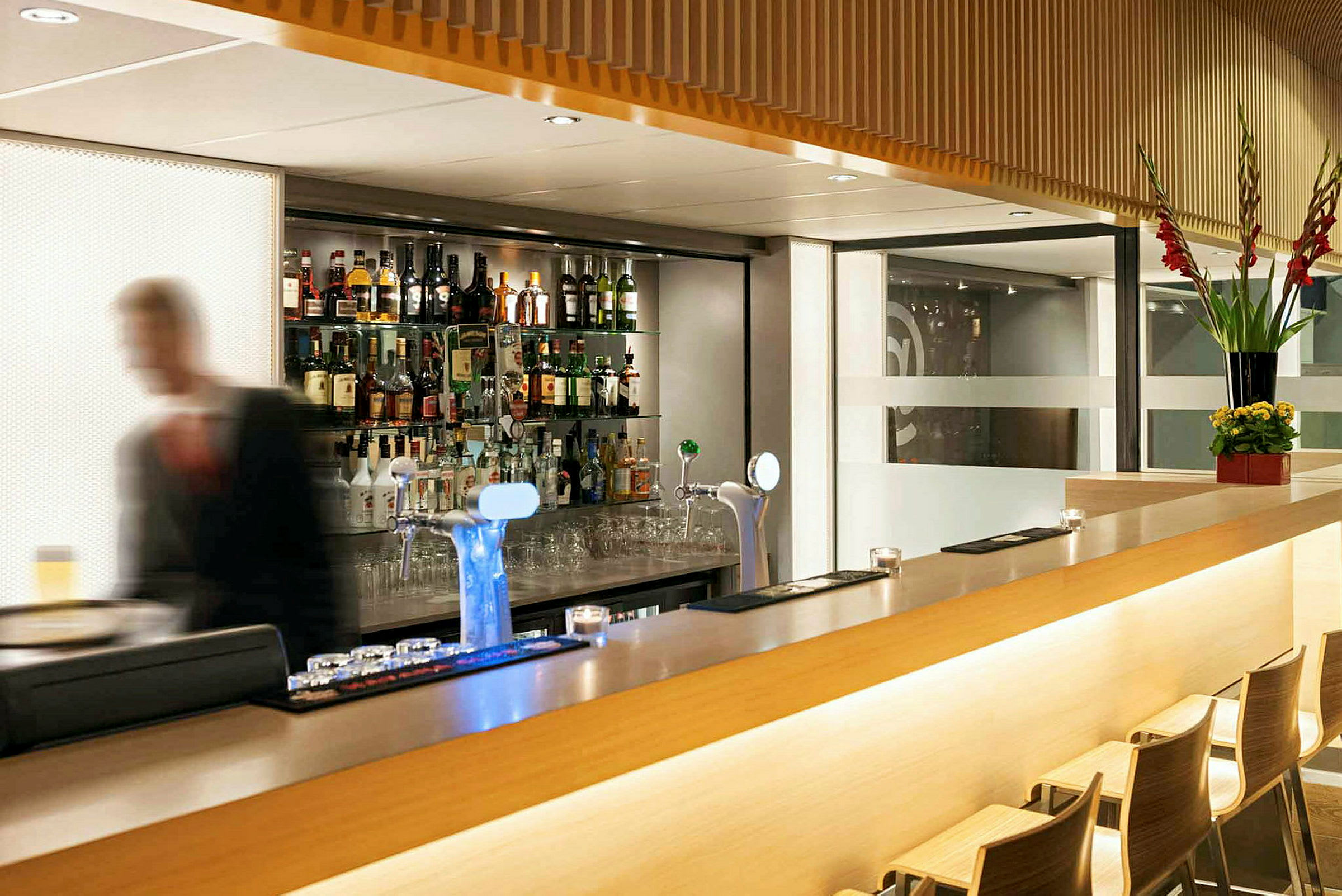 En modern bar med ett utbud av flaskor på hyllor bakom samt en blurrad person i förgrunden. (AI-genererad text)