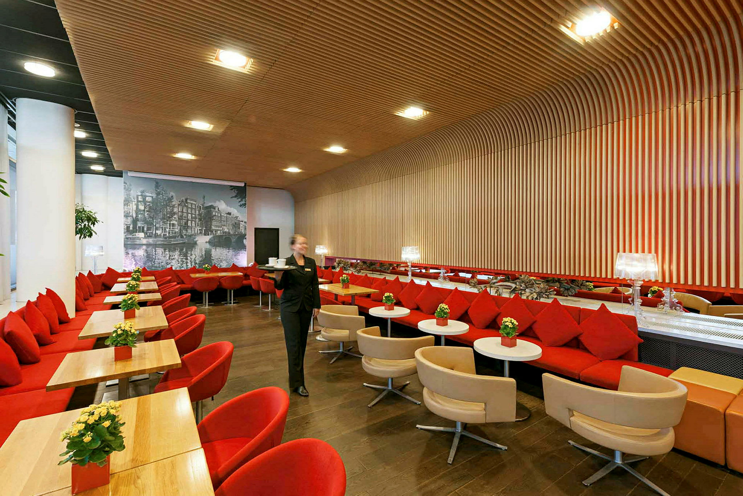 En restaurang med moderna inslag, röda och beige sittplatser, en stor vägg med träpaneler och en bild på staden i bakgrunden. Bord och stolar är placerade i rader. (AI-genererad text)