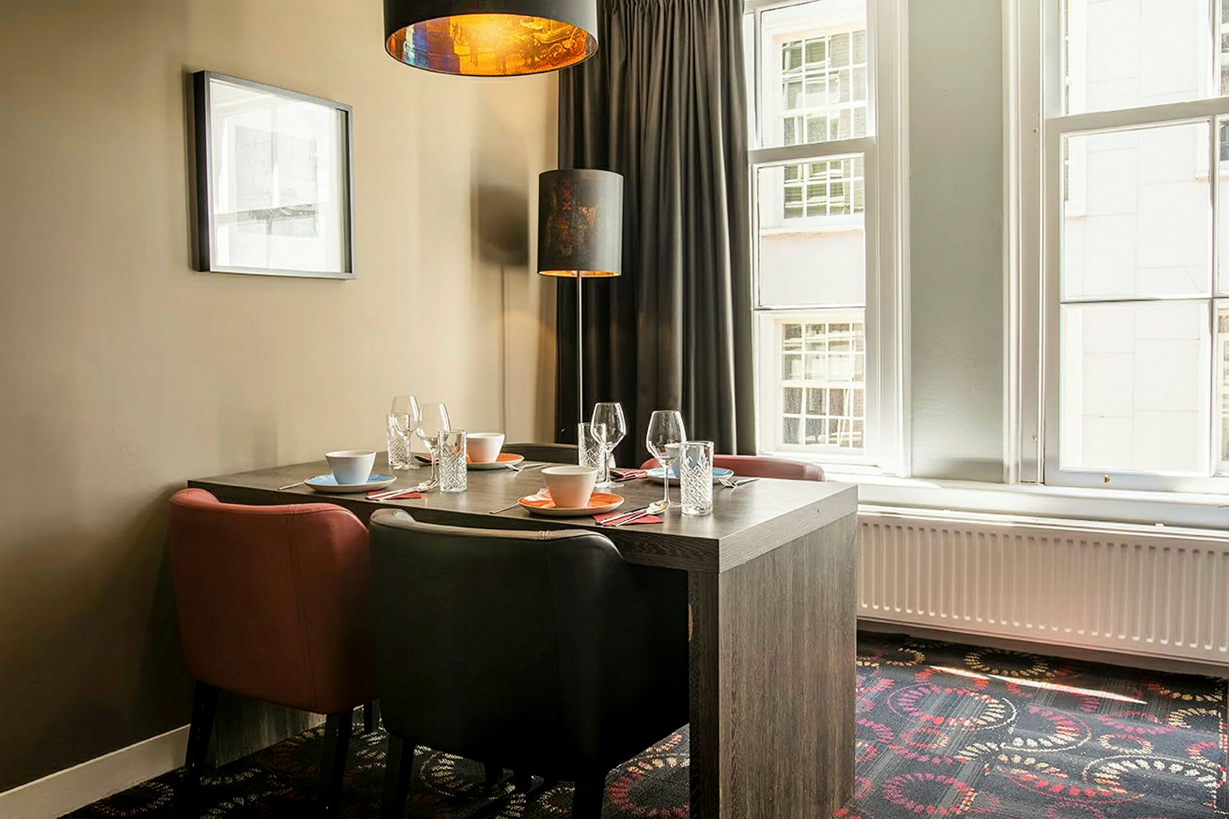 Ett matbord med fyra stolar. Bordet är dukat med glas, tallrikar och skålar. En orange hängande lampa lyser över bordet. Fönster och gardiner syns i bakgrunden. (AI-genererad text)