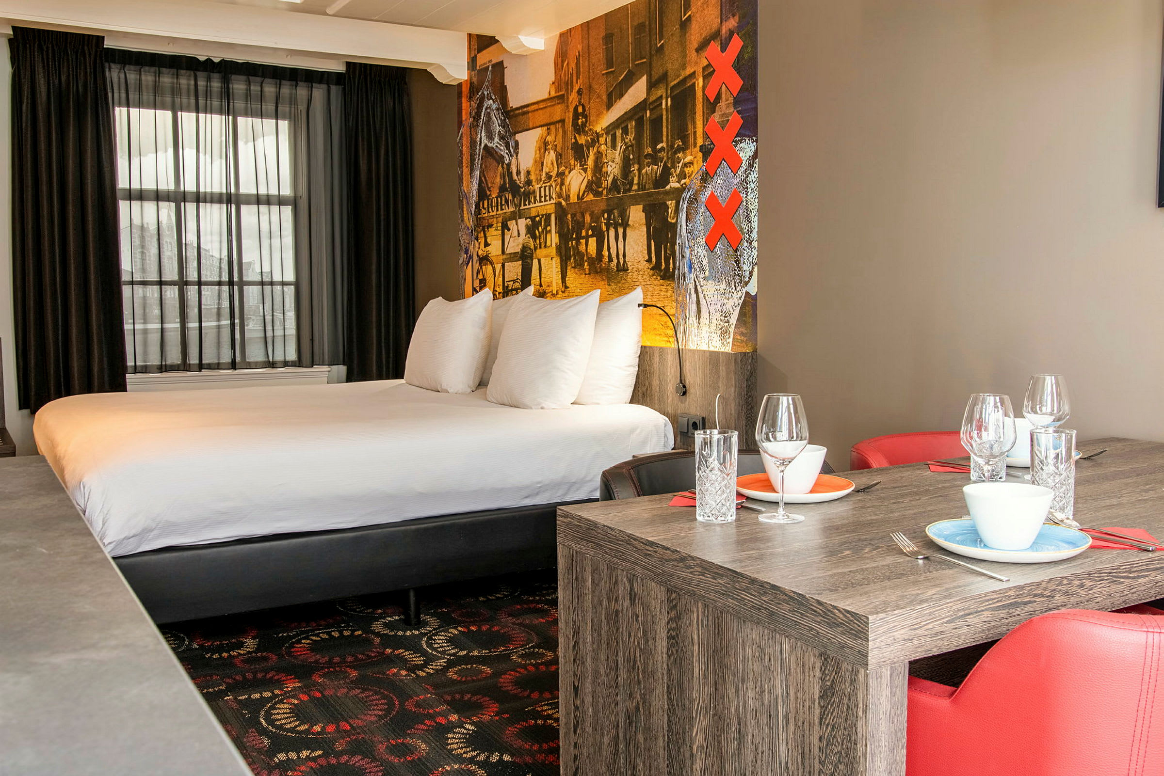 Ett hotellrum med en stor säng, röda stolar och ett matbord med dukade tallrikar och glas. (AI-genererad text)