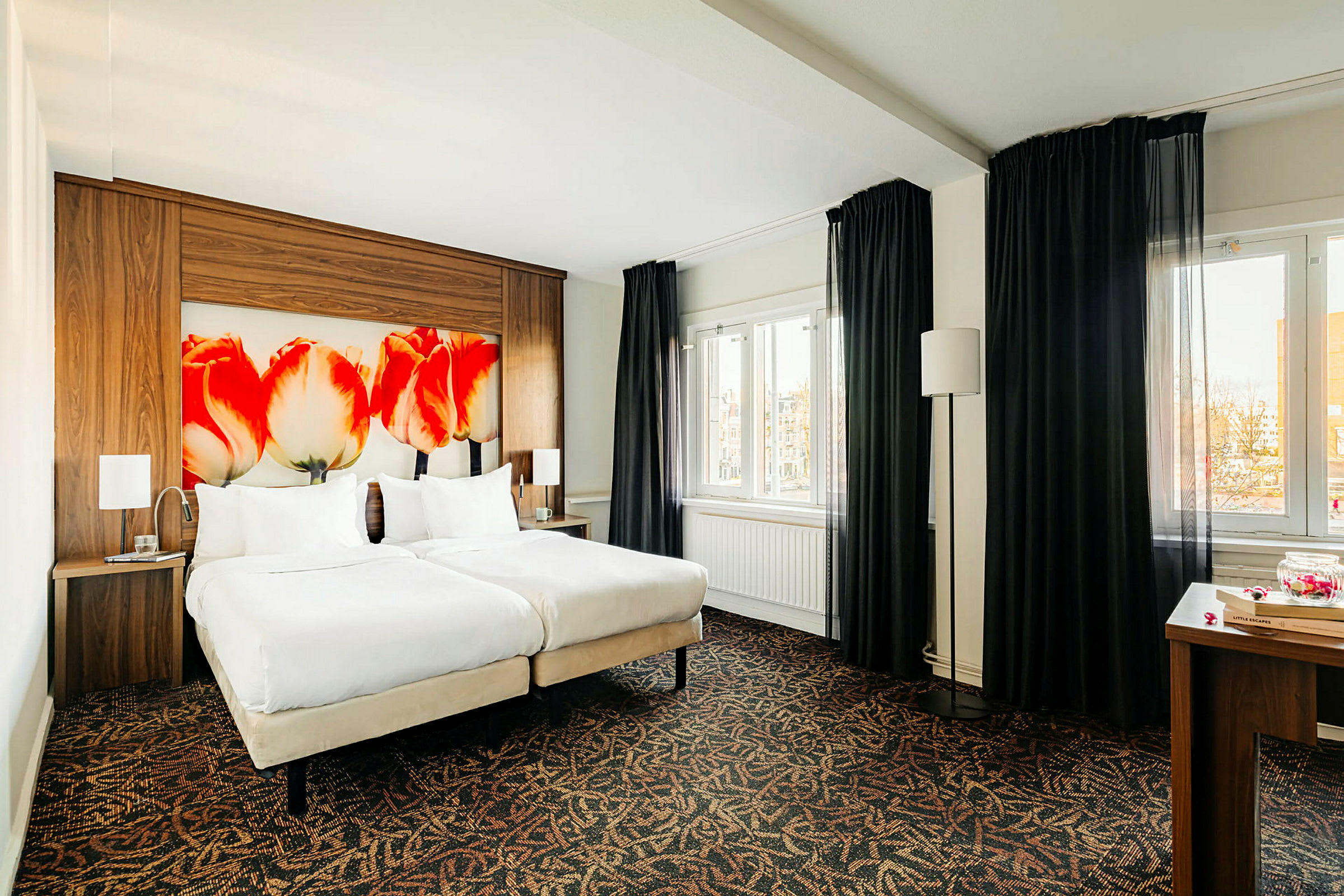 Ett hotellrum med två enkelsängar och en sänggavel med tulpanmotiv. Det finns mörkläggningsgardiner vid fönstren. (AI-genererad text)
