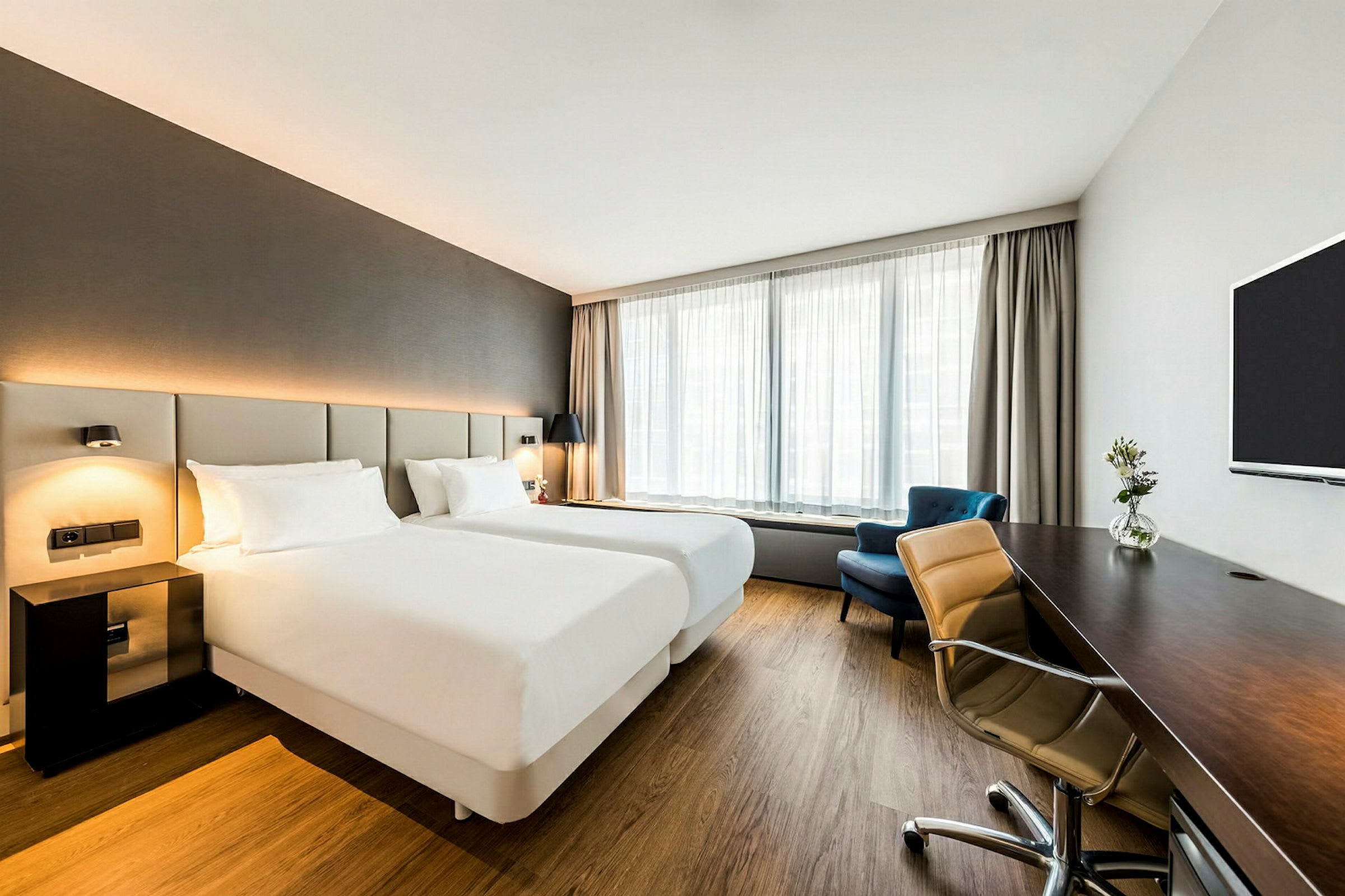 Ett ljust hotellrum med två enkelsängar, ett skrivbord, en tv och ett fönster med gardiner. (AI-genererad text)
