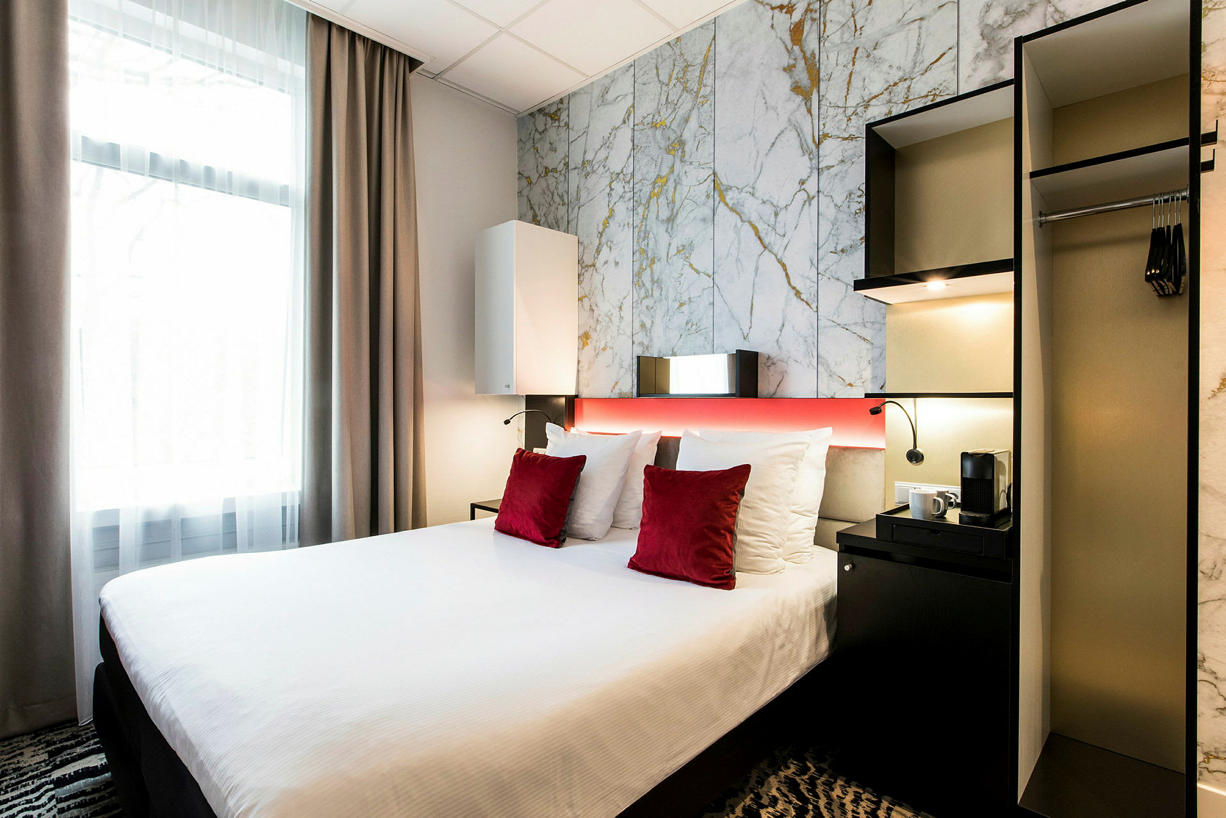 Ett hotellrum med en säng med vita sängkläder och röda prydnadskuddar, bredvid en garderob och ett litet bord med en kaffemaskin. (AI-genererad text)