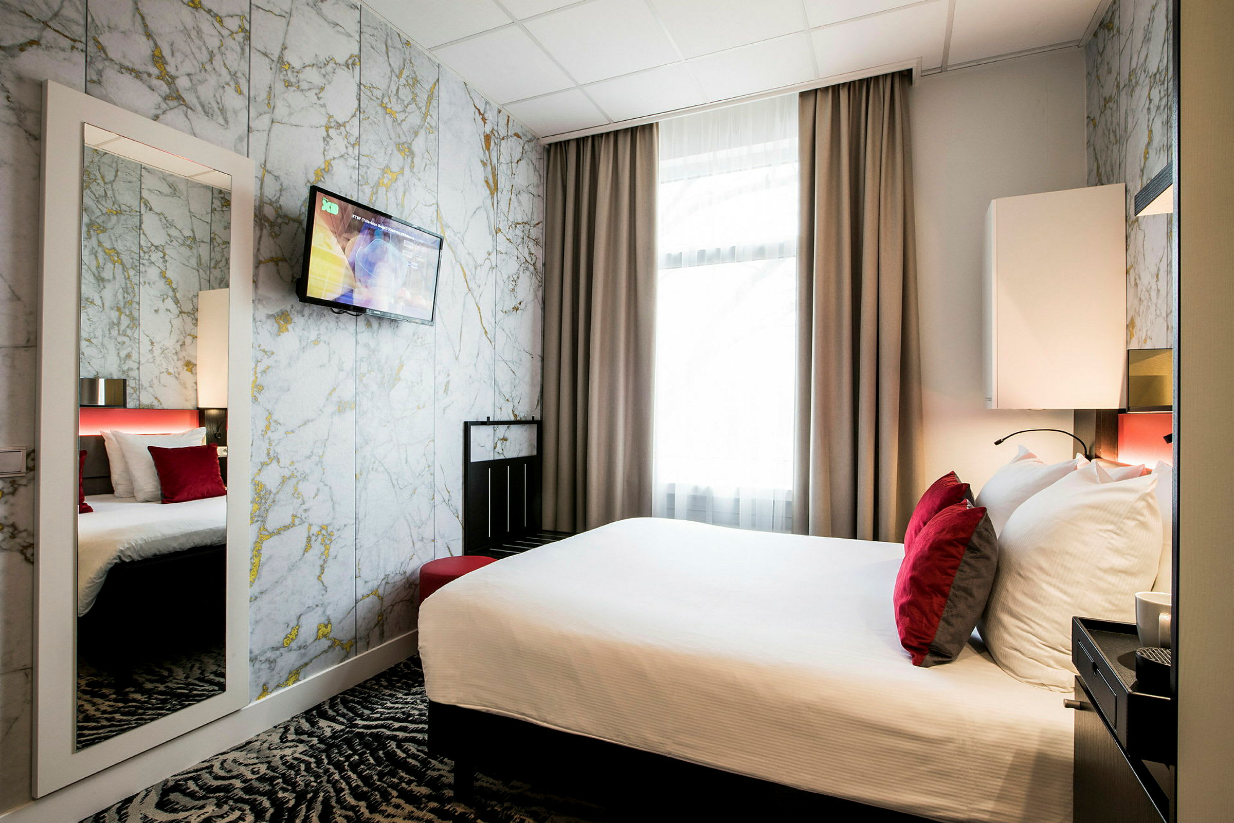 Ett hotellrum med en dubbelsäng med vita lakan och röda kuddar framför ett marmorliknande väggmönster och en väggmonterad TV. (AI-genererad text)
