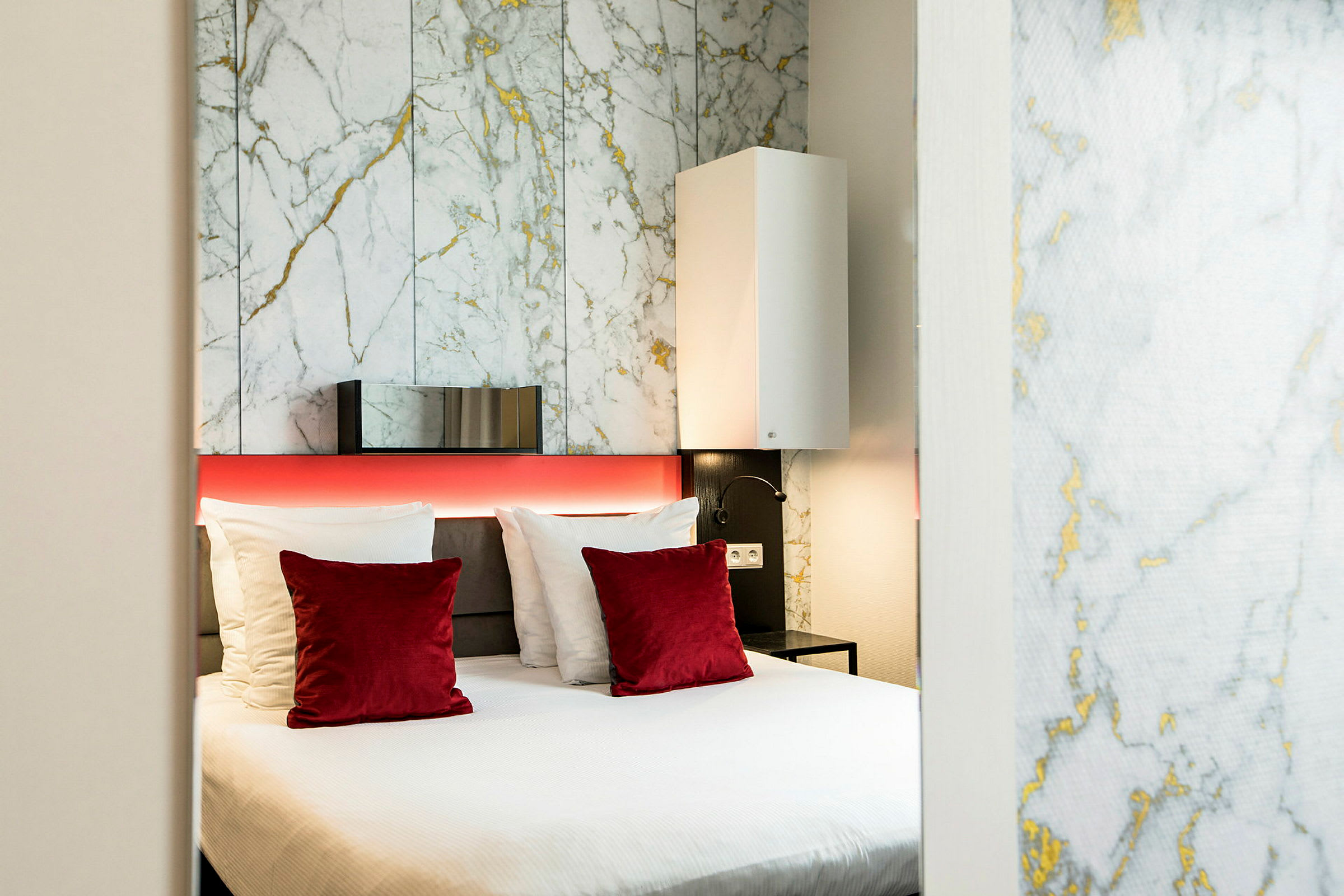Ett modernt hotellrum med en dubbelsäng, vita och röda kuddar samt en bakgrundsvägg med marmormönster. (AI-genererad text)