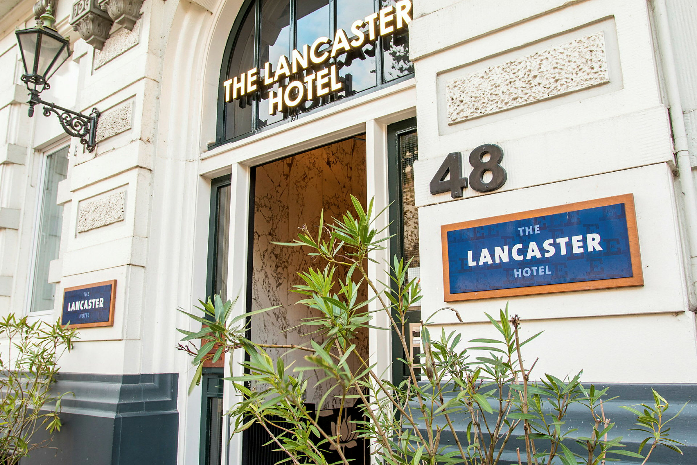 Entré till ett hotell med namnet 'The Lancaster Hotel', pryds av gröna växter. (AI-genererad text)