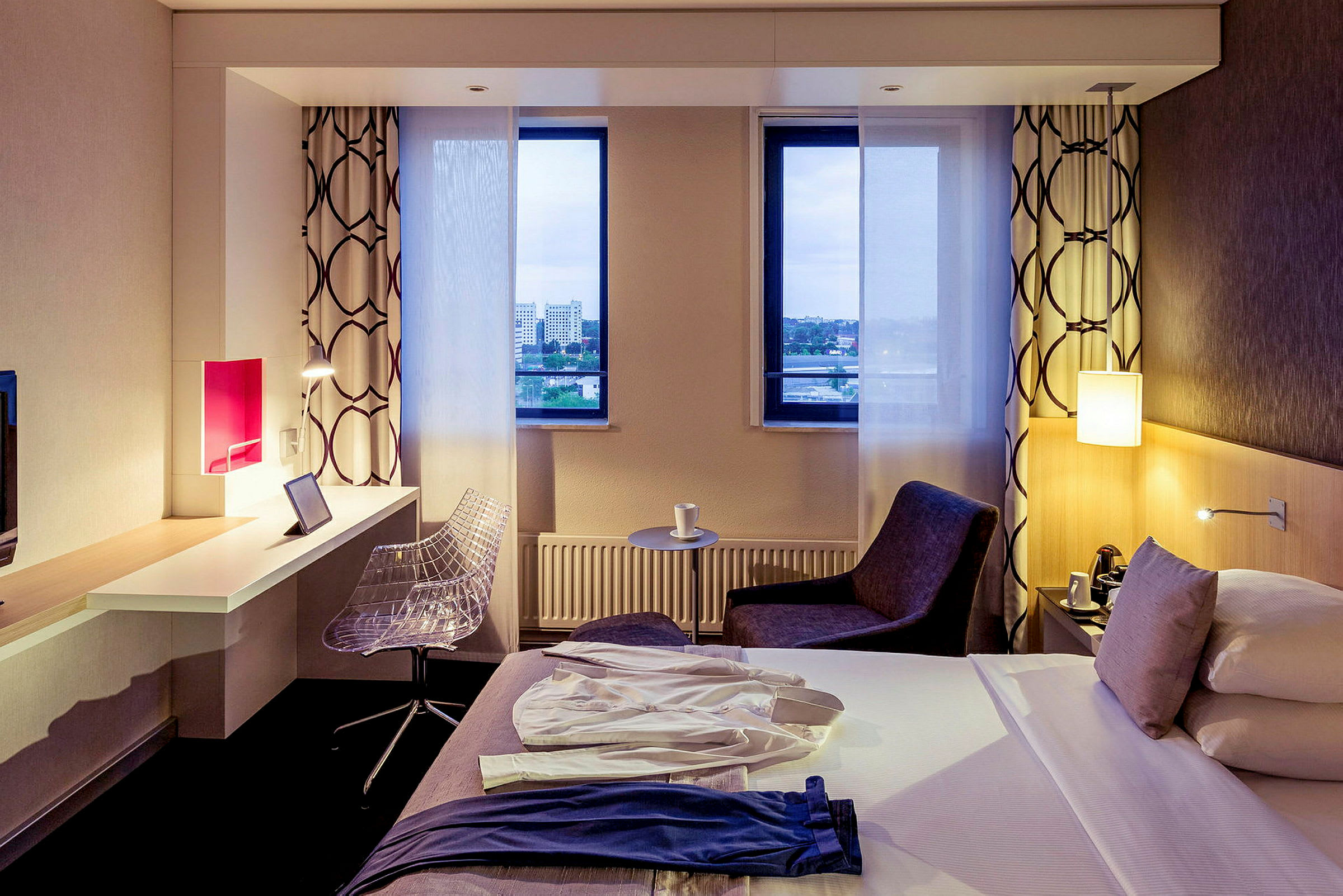 Ett hotellrum med en säng, skrivbord och stol samt två fönster med utsikt. (AI-genererad text)