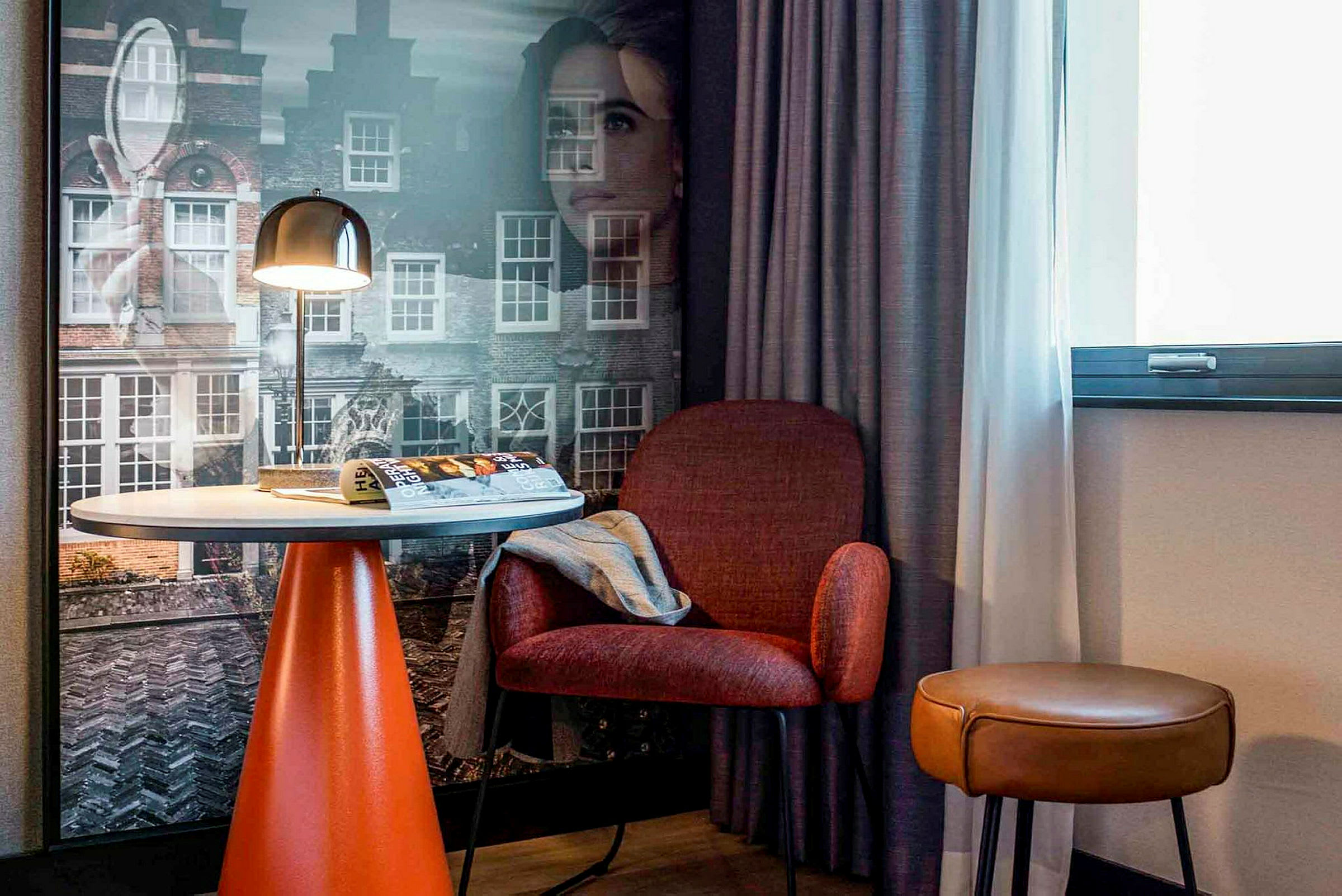 Ett hotellrum med ett bord, en röd stol och en brun pall framför ett fönster och en vägg med stadsmotiv. (AI-genererad text)