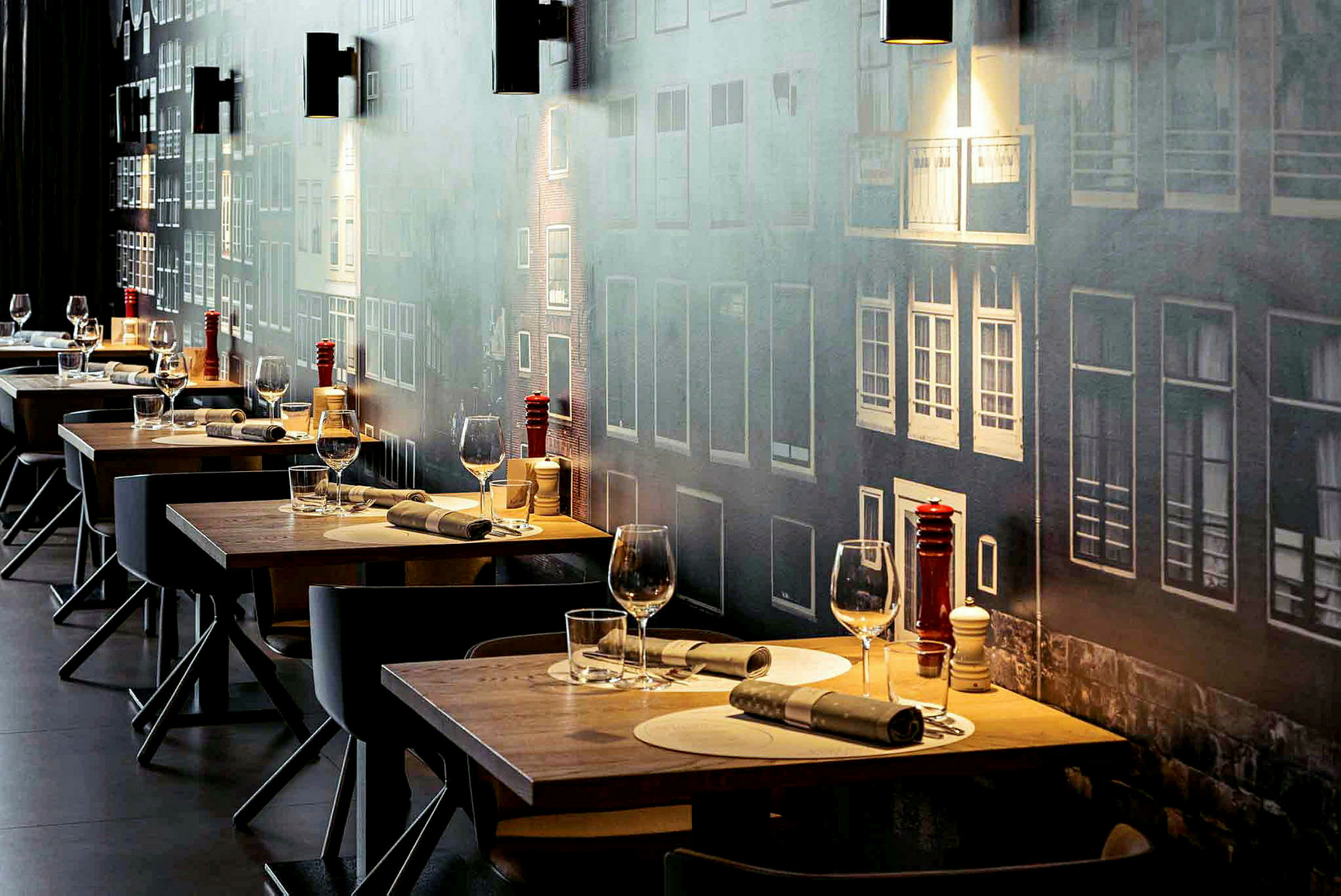 Ett antal dukade restaurangbord med glas och bestick, mot en vägg med fasadmotiv. (AI-genererad text)