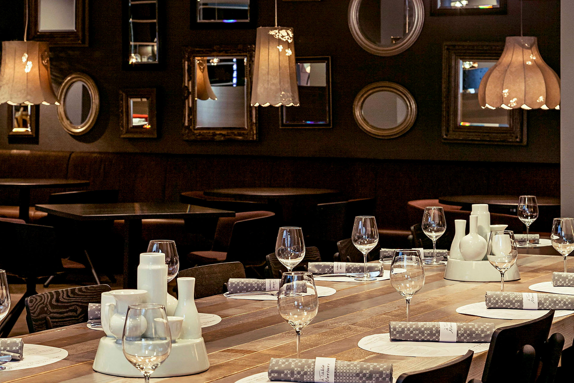 En restaurang med dukade bord och dekorationer på en träyta, omgiven av mörka väggar med speglar och lampor. (AI-genererad text)