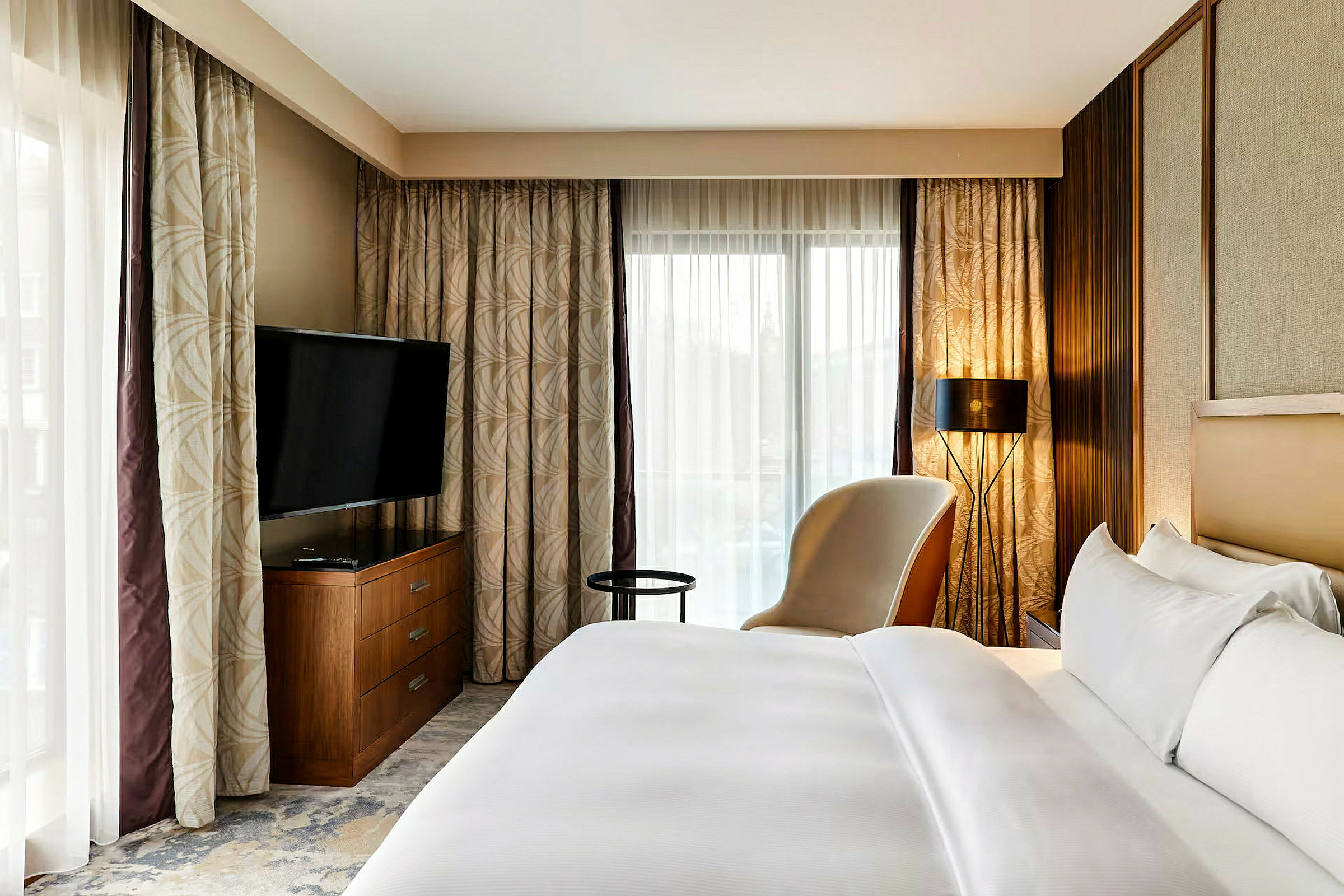 Ett hotellrum med en bekväm säng, en fåtölj, ett sidobord, en TV på en byrå och fönster med gardiner. (AI-genererad text)