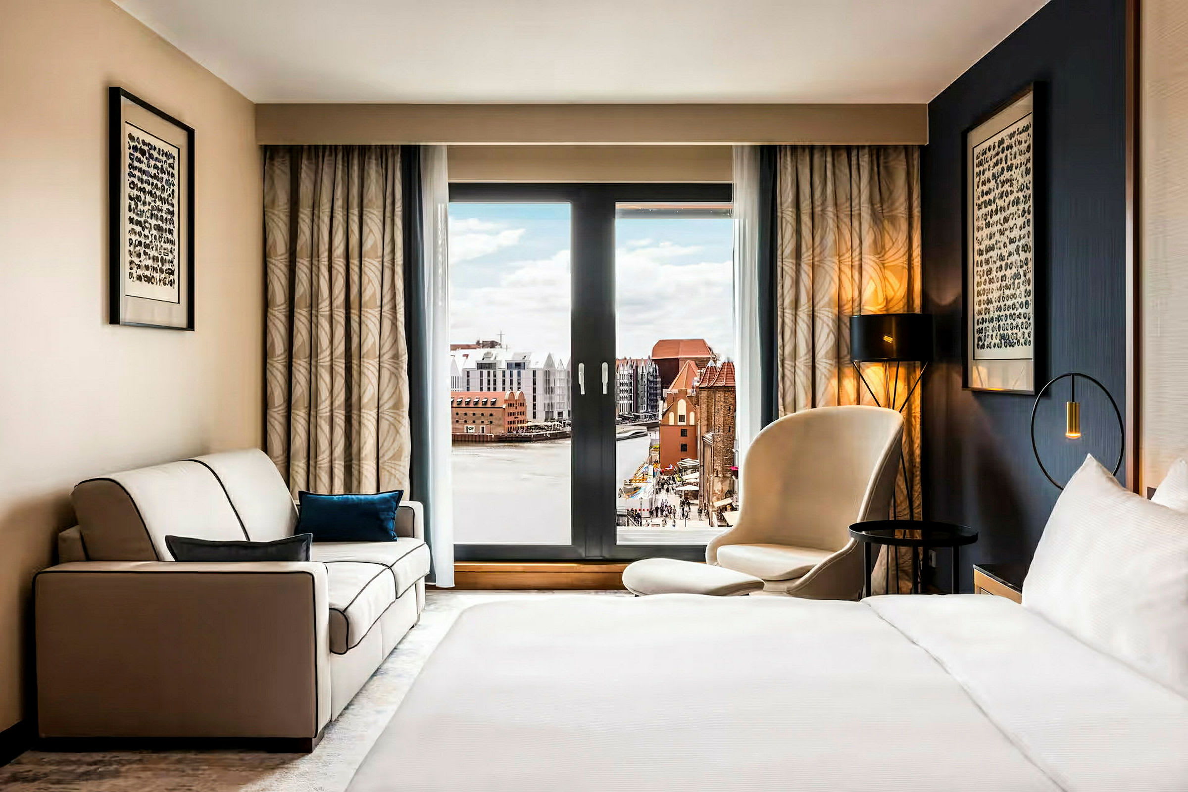 Ett hotellrum med en säng, en soffa och en stol framför ett stort fönster med utsikt över en stad och vatten. (AI-genererad text)